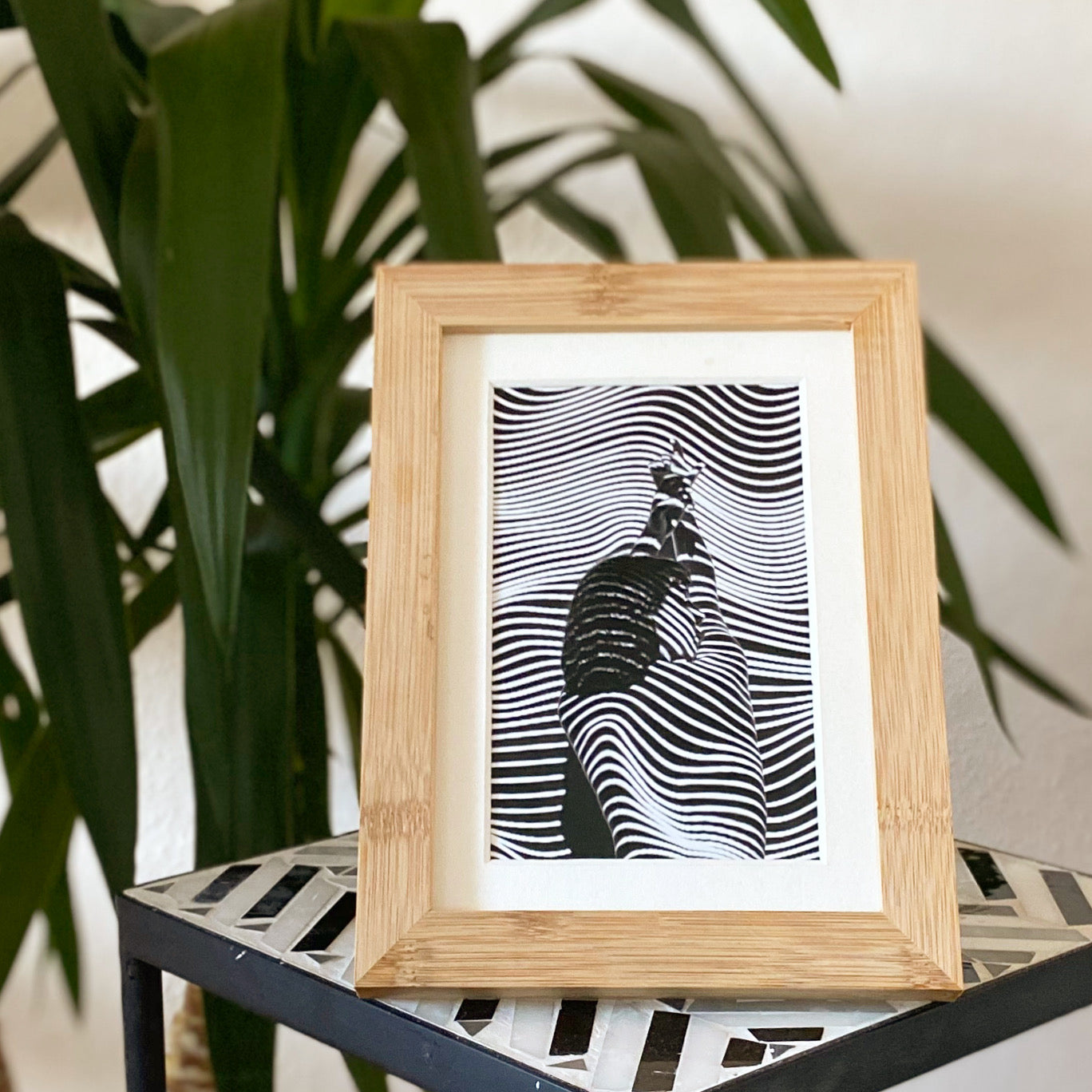 Holzrahmen mit abstraktem, schwarz-weißem Kunstwerk, das optische Linien zeigt; im Hintergrund grüne Pflanzen.