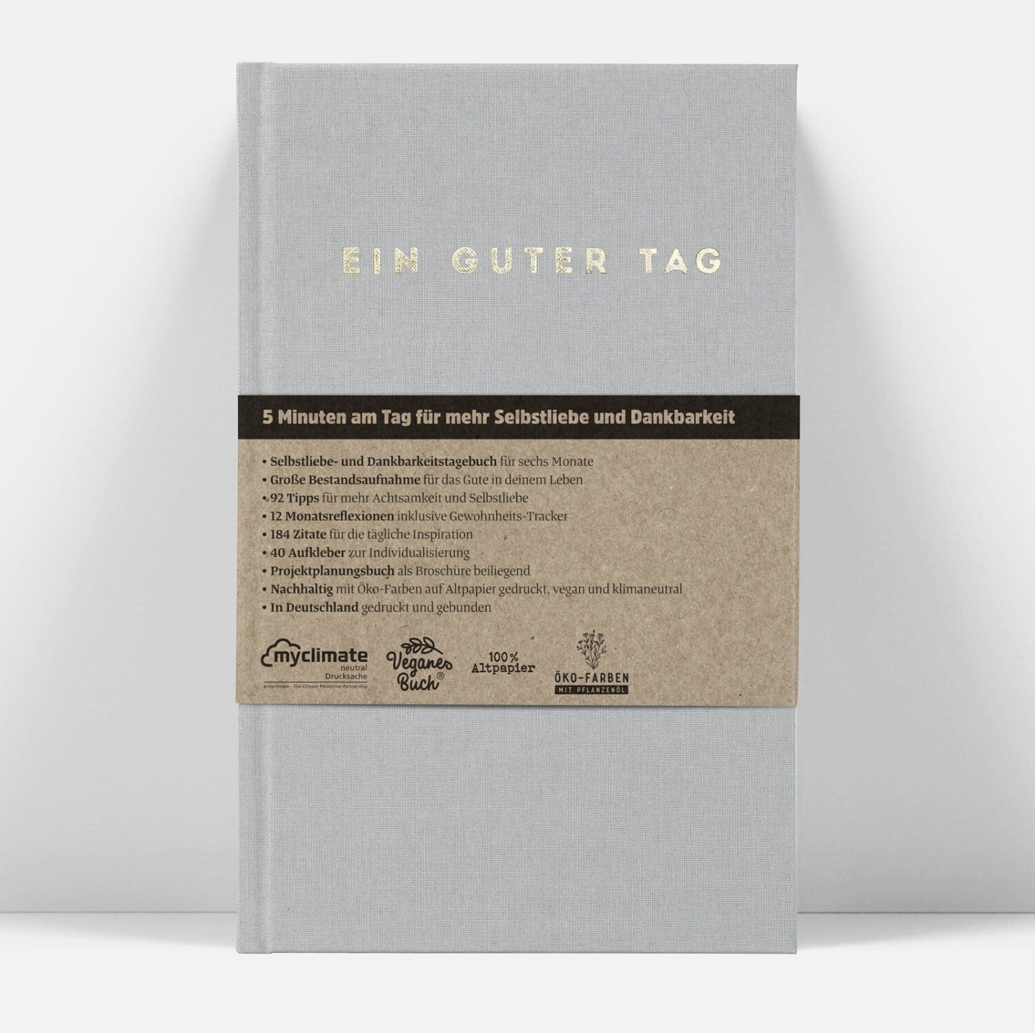 Graues Notizbuch mit der Aufschrift "EIN GUTER TAG" und beige Banderole mit Detailtext zu Inhalten und Nachhaltigkeit.