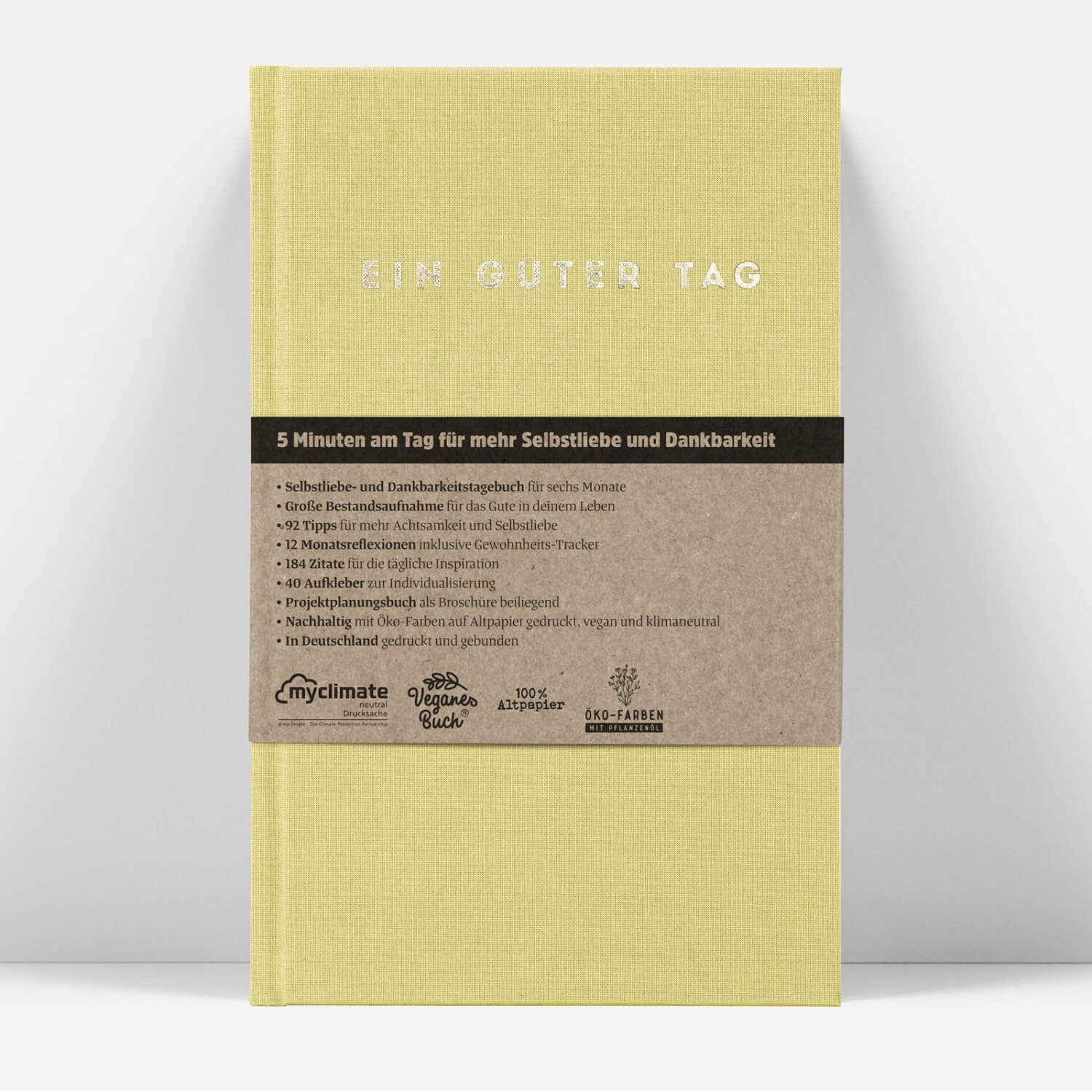 Ein gelbes Tagebuch mit der Aufschrift "Ein guter Tag" und einem braunen Band, das Details zu Inhalt und Nachhaltigkeit erklärt.