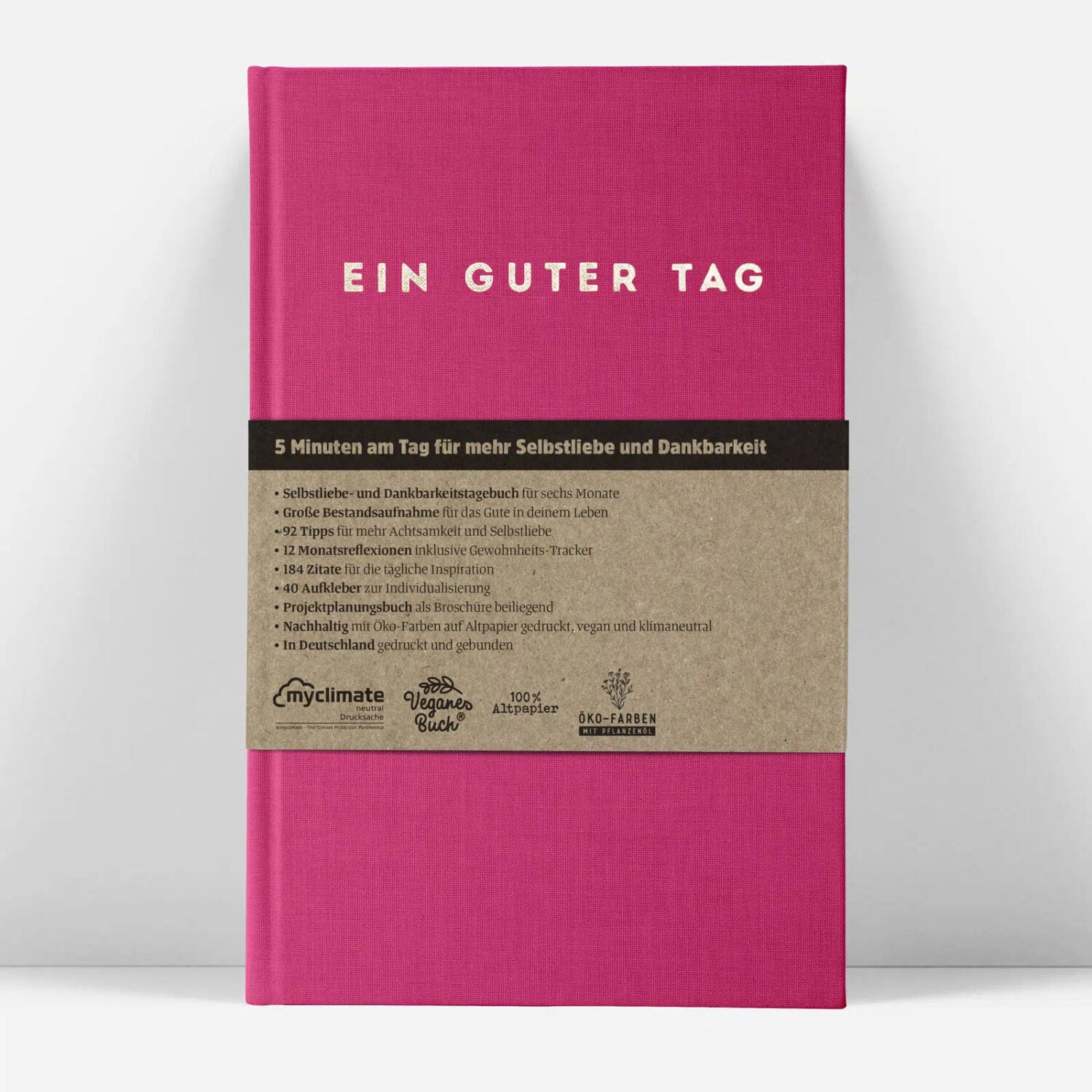 Ein pinkes Buch mit dem Titel "EIN GUTER TAG" und Details zu Selbstliebe und Dankbarkeit auf einem braunen Umschlag.