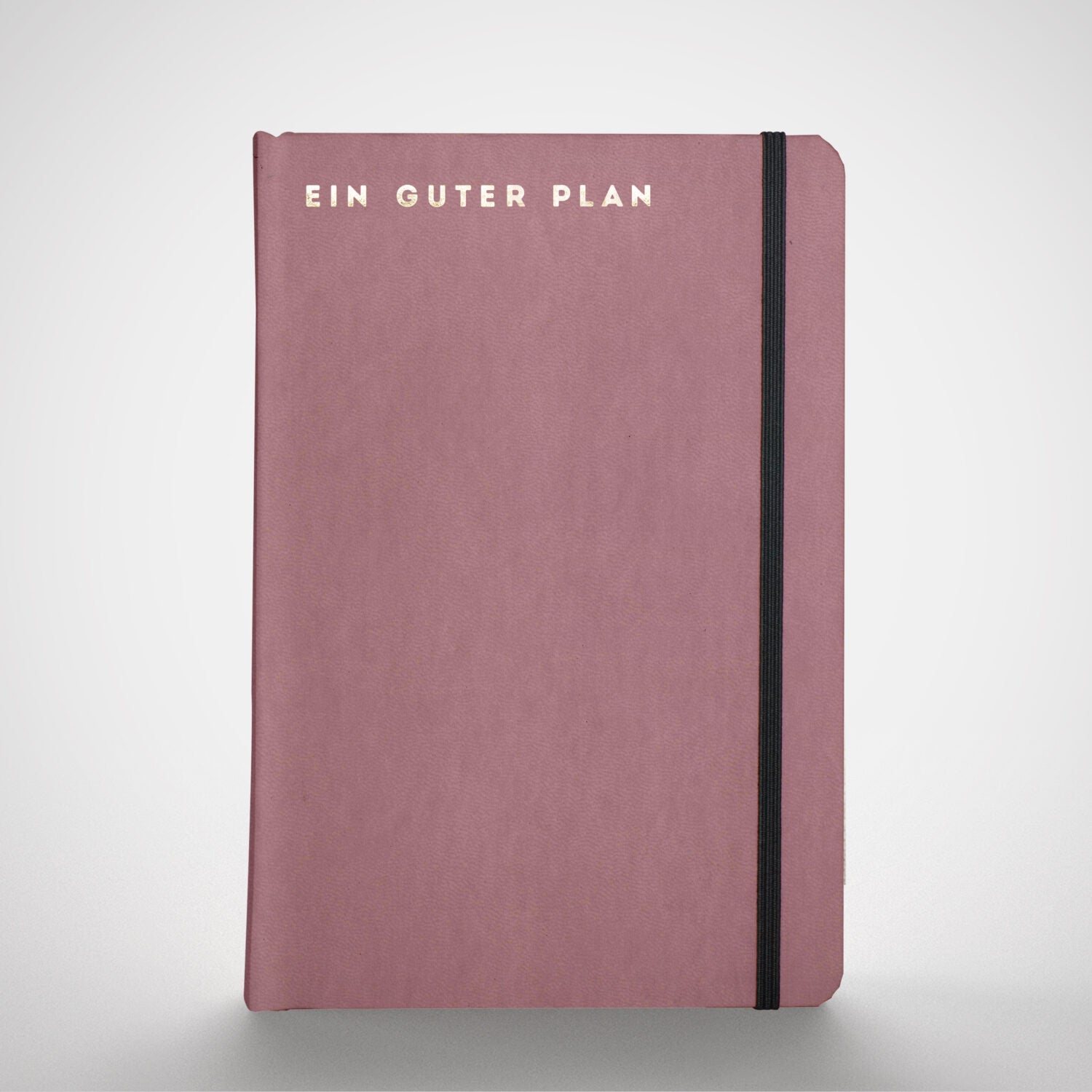 Rosa Notizbuch mit der Aufschrift "EIN GUTER PLAN" in Gold und einem schwarzen Gummiband an der rechten Seite.