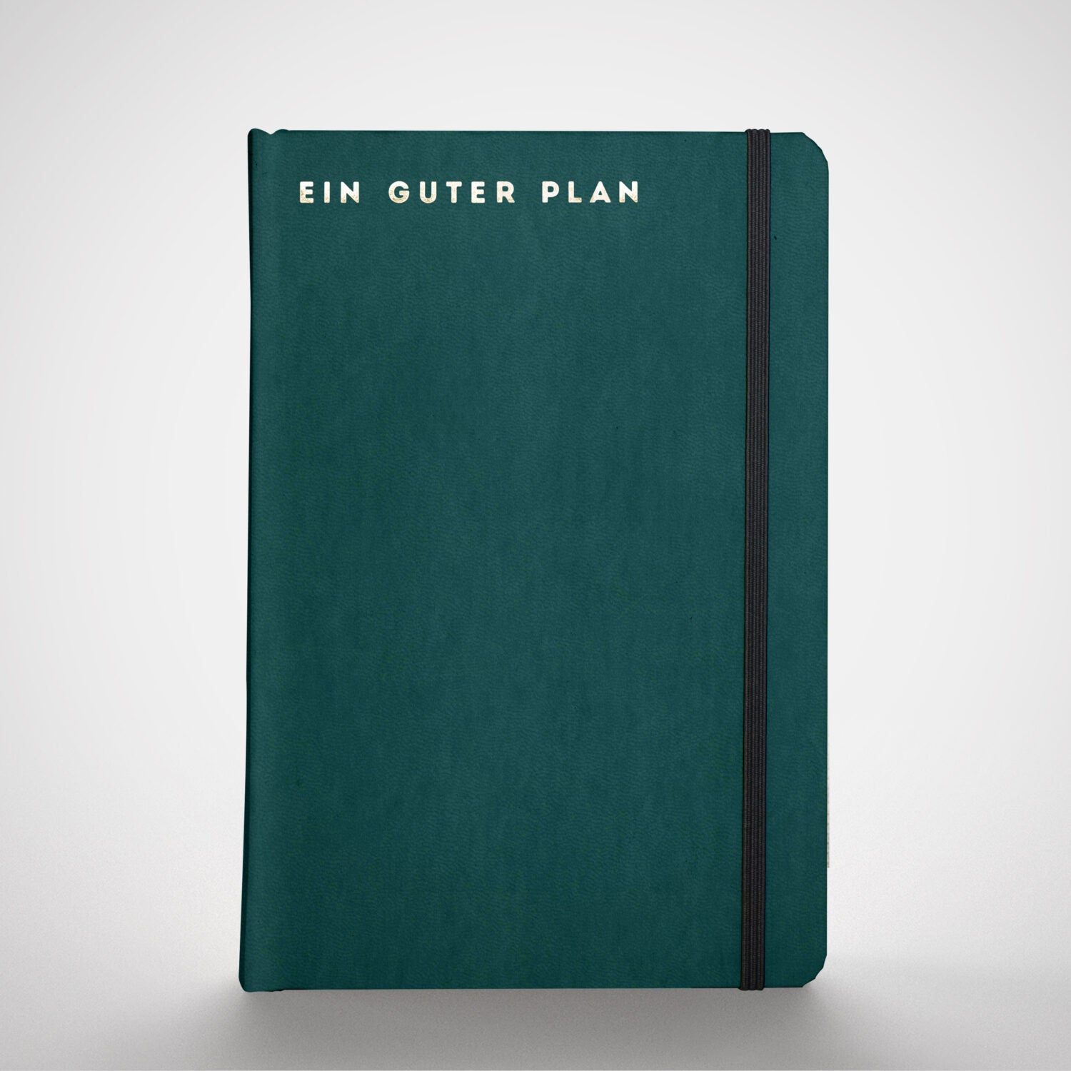 Grünes Notizbuch mit der Aufschrift "Ein Guter Plan" und einem schwarzen Gummiband auf grauem Hintergrund.