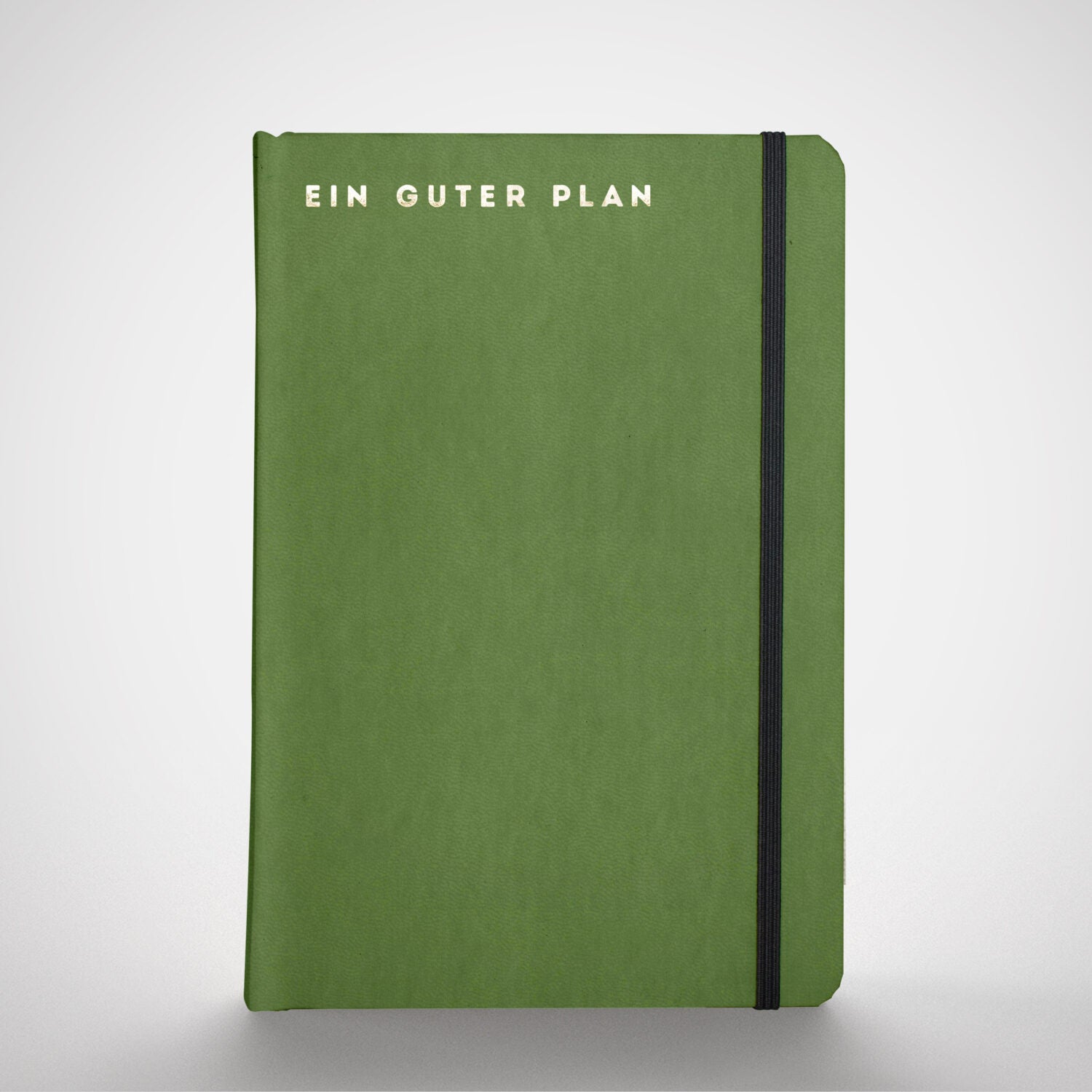 Grünes Notizbuch mit der Aufschrift "Ein Guter Plan" und einem schwarzen Verschlussband auf grauem Hintergrund.