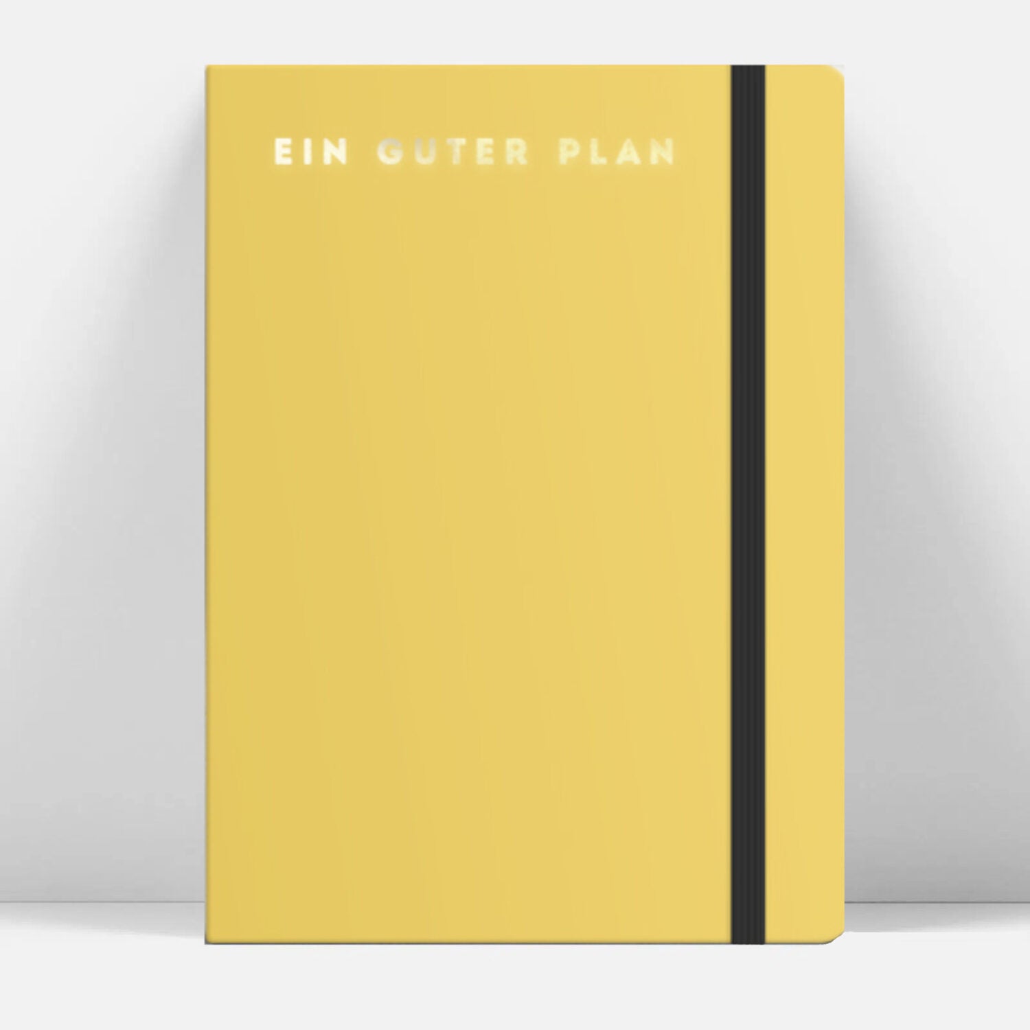 Gelbes Notizbuch mit weißem Schriftzug "EIN GUTER PLAN" und schwarzem Gummiband auf hellem Hintergrund.