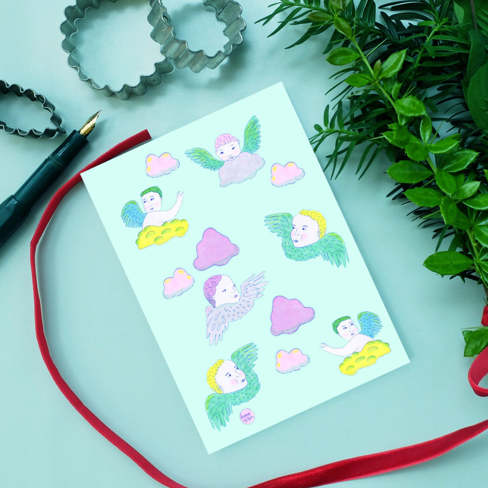 Eine Karte mit Illustrationen von Engeln und Wolken, umgeben von Keksausstechern, einem Stift und Pflanzen.