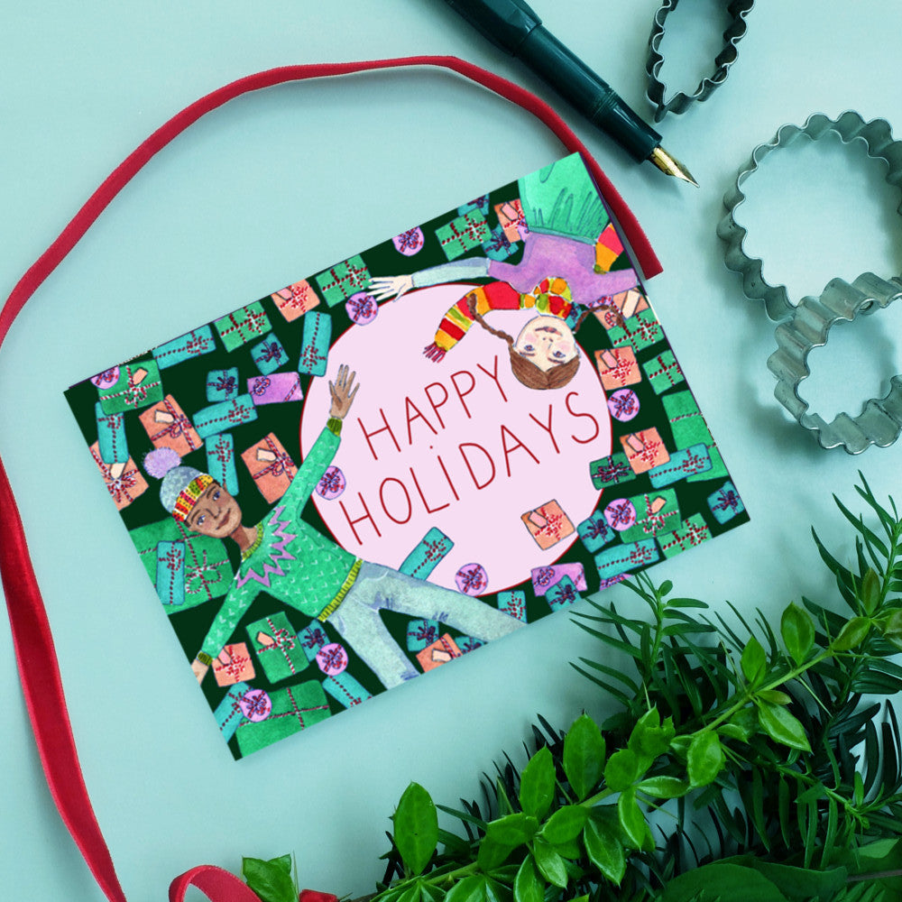 Weihnachtskarte mit "Happy Holidays" und zwei gezeichneten Figuren, umgeben von Geschenken, neben Keksausstechern und Grün.