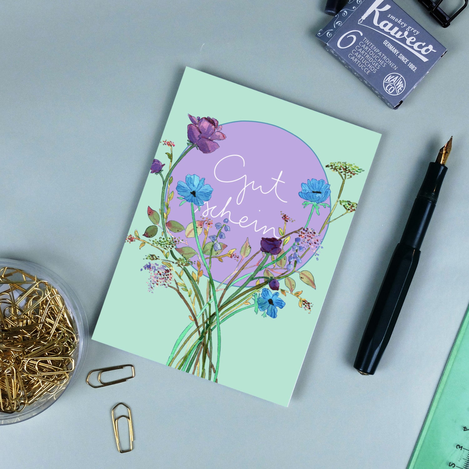 Eine Geschenkkarte mit Blumenmotiven und der Aufschrift "Gutschein" liegt auf einem Schreibtisch mit Büroklammern und Stift.
