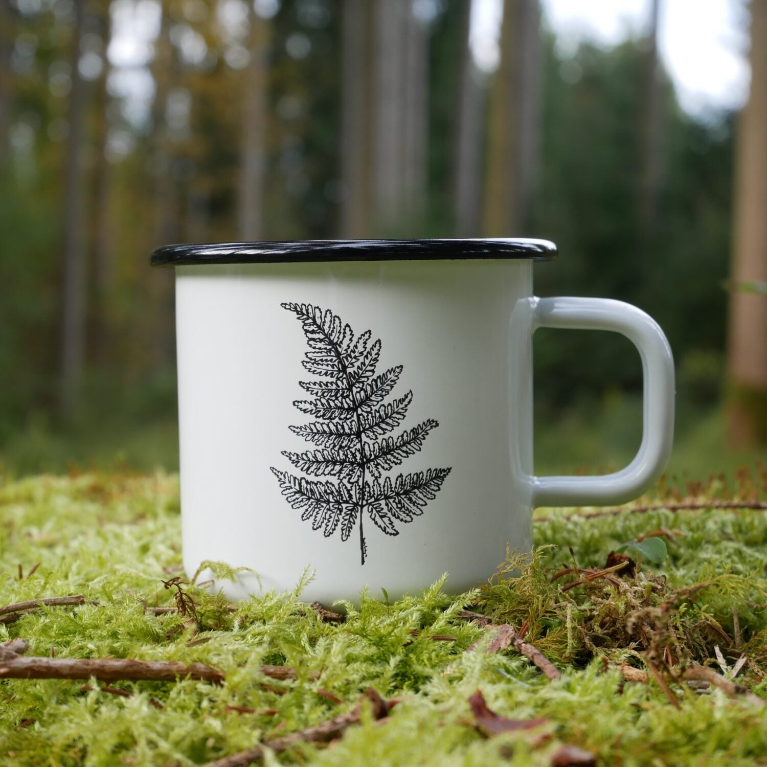 Eine weiße Tasse mit Farnmuster steht auf moosbedecktem Boden im Wald.