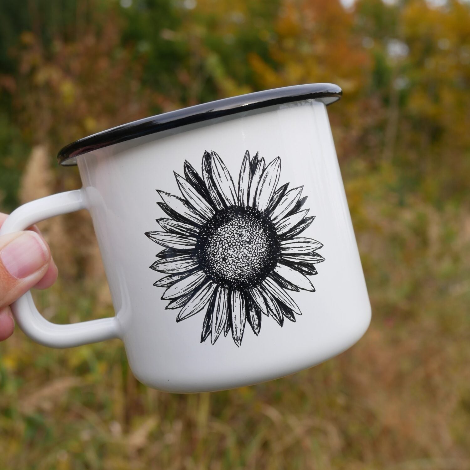 Weiße Emaille-Tasse mit Sonnenblumen-Motiv, von einer Hand vor herbstlicher Naturkulisse gehalten.