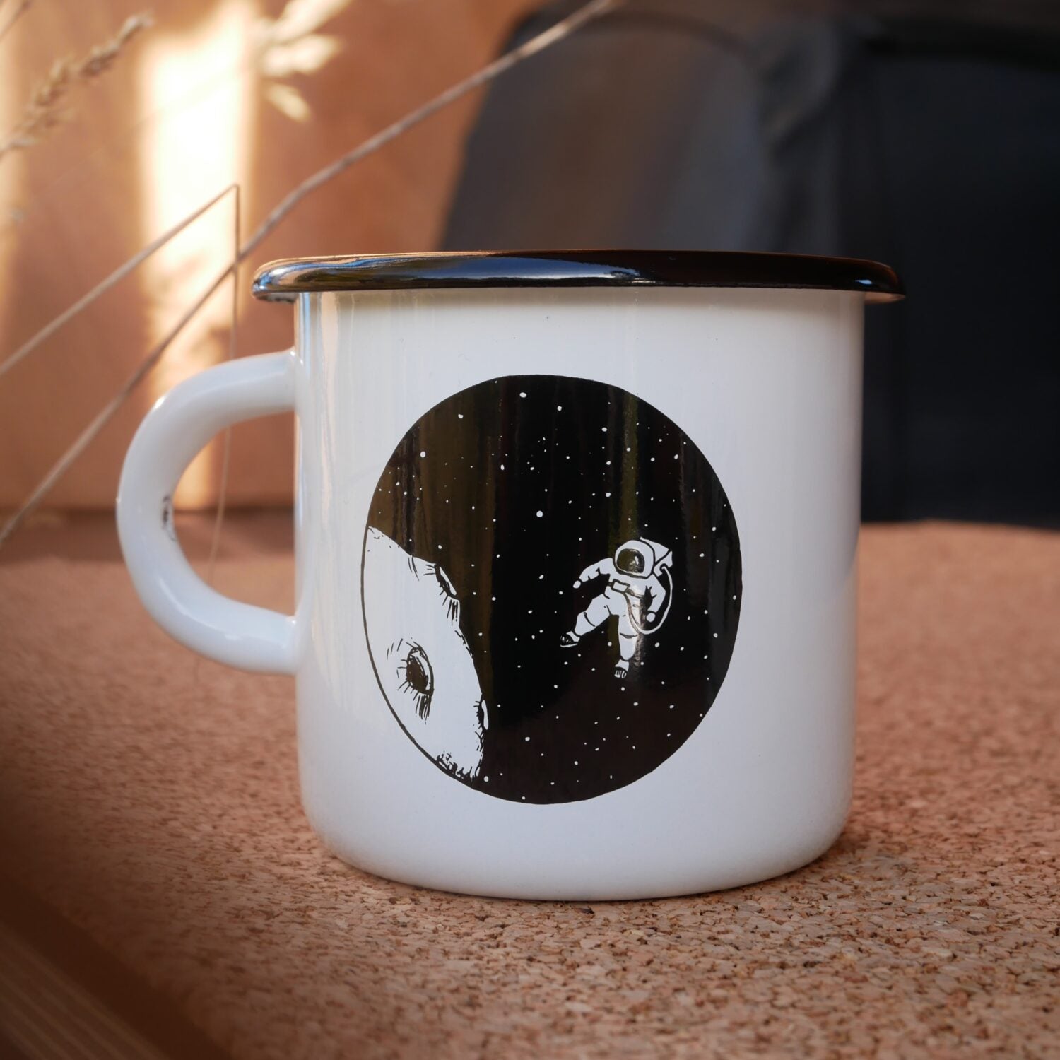 Weiße Emaille-Tasse mit einem Astronauten und Mond-Illustration auf einer Korkunterlage.