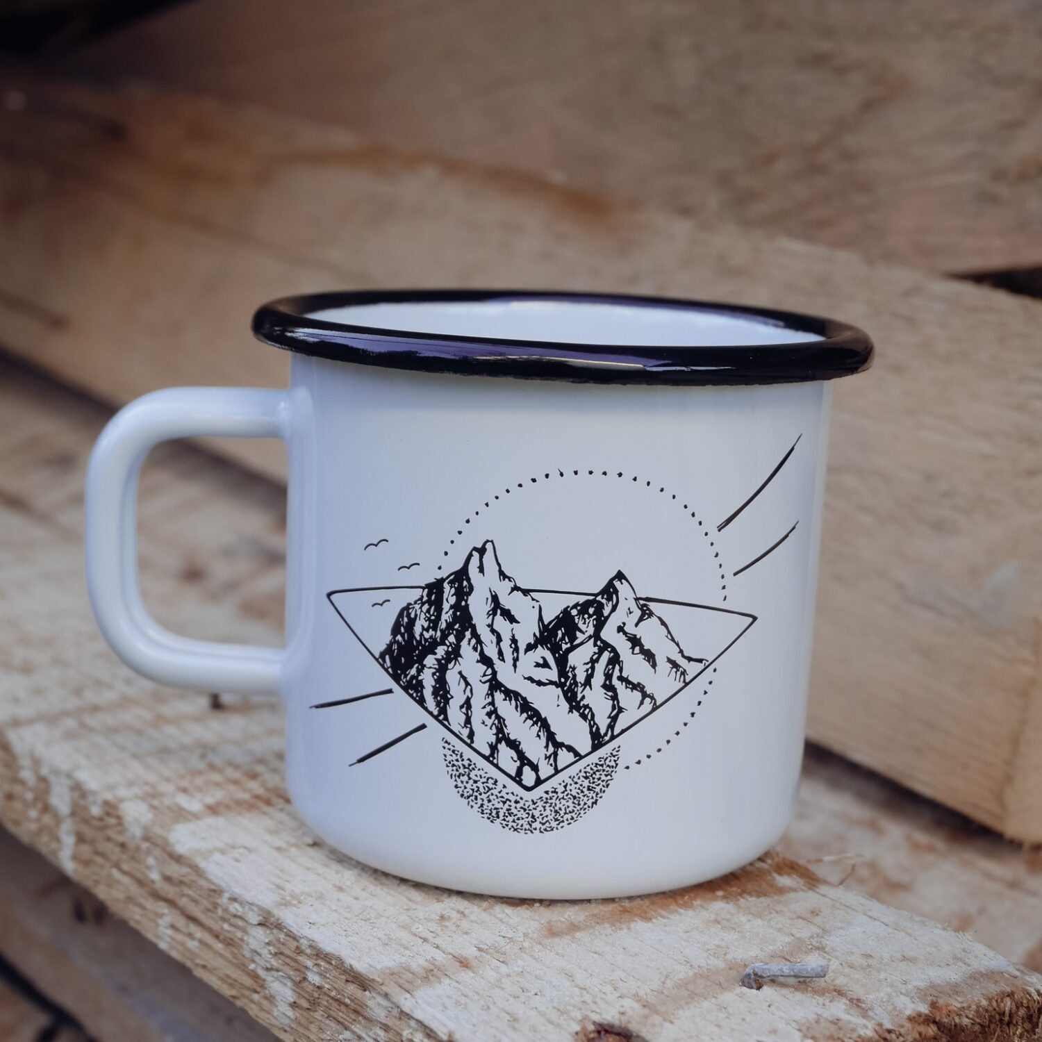 Weiße Emaille-Tasse mit schwarzem Rand und einem Bergmotiv, auf einem hölzernen Untergrund stehend.