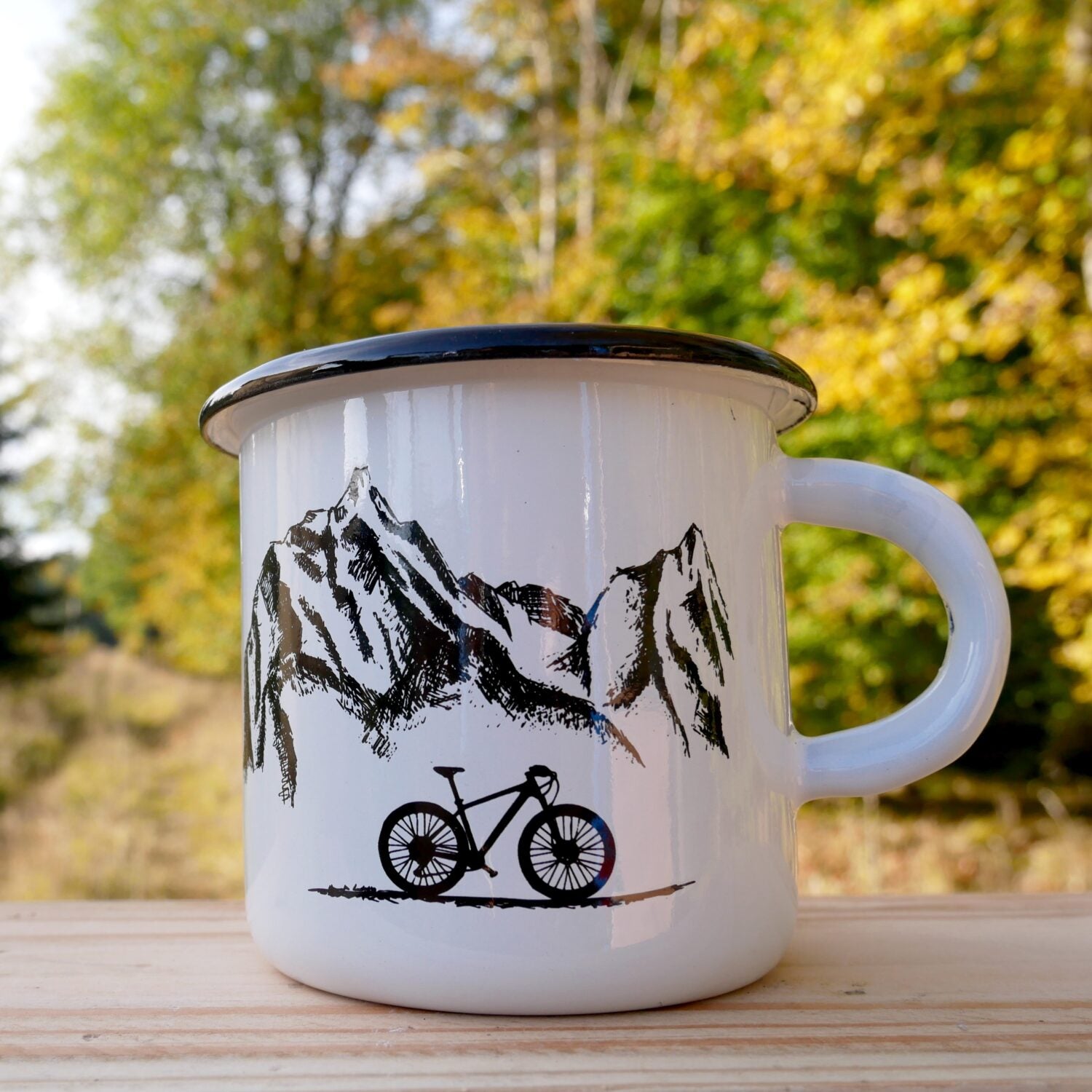 Weiße Tasse mit Mountainbike- und Bergmuster, steht auf Holzuntergrund vor einem herbstlichen Waldhintergrund.