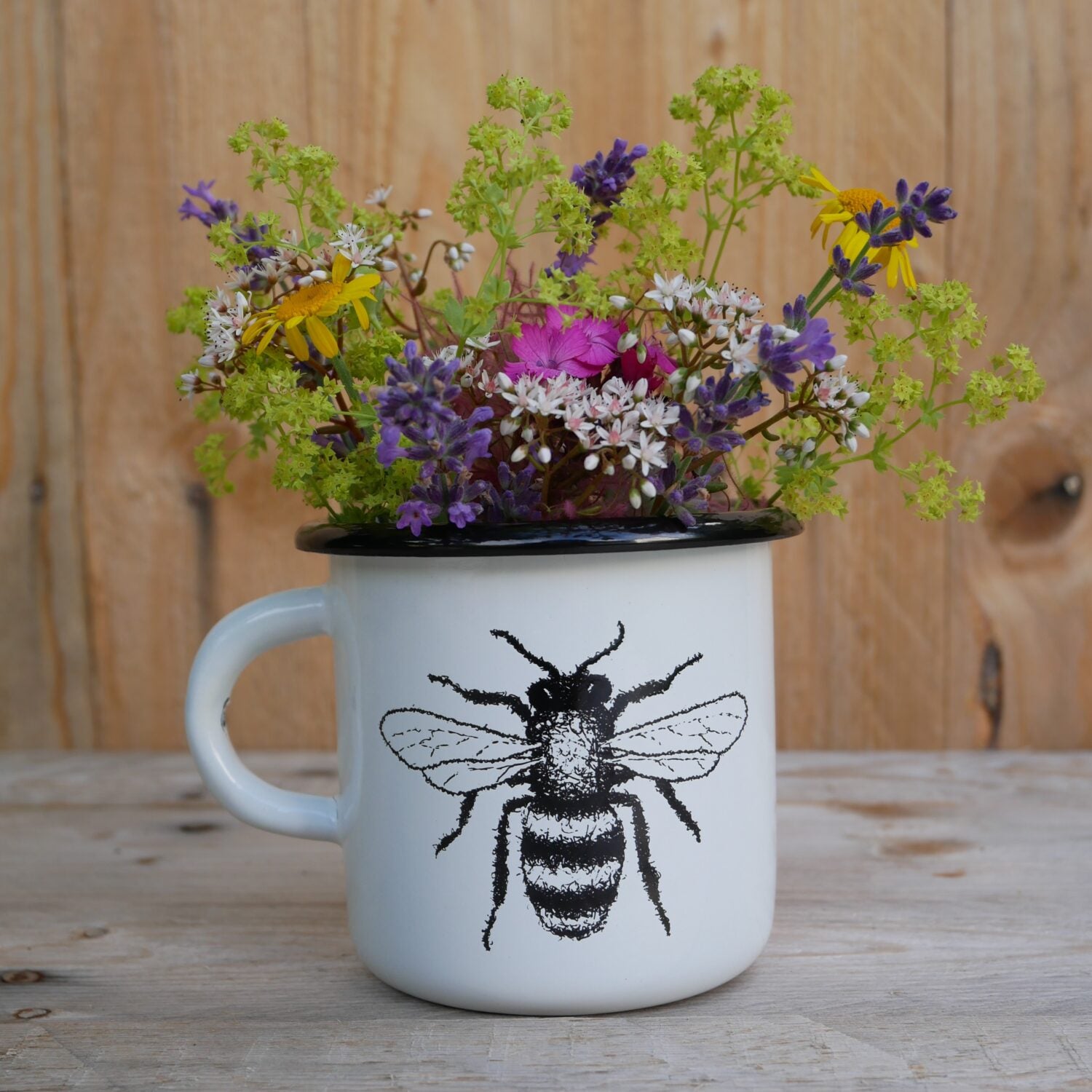 Weiße Tasse mit Bienenmotiv, gefüllt mit bunten Wildblumen, steht auf einem Holztisch vor einer Holzplatte.