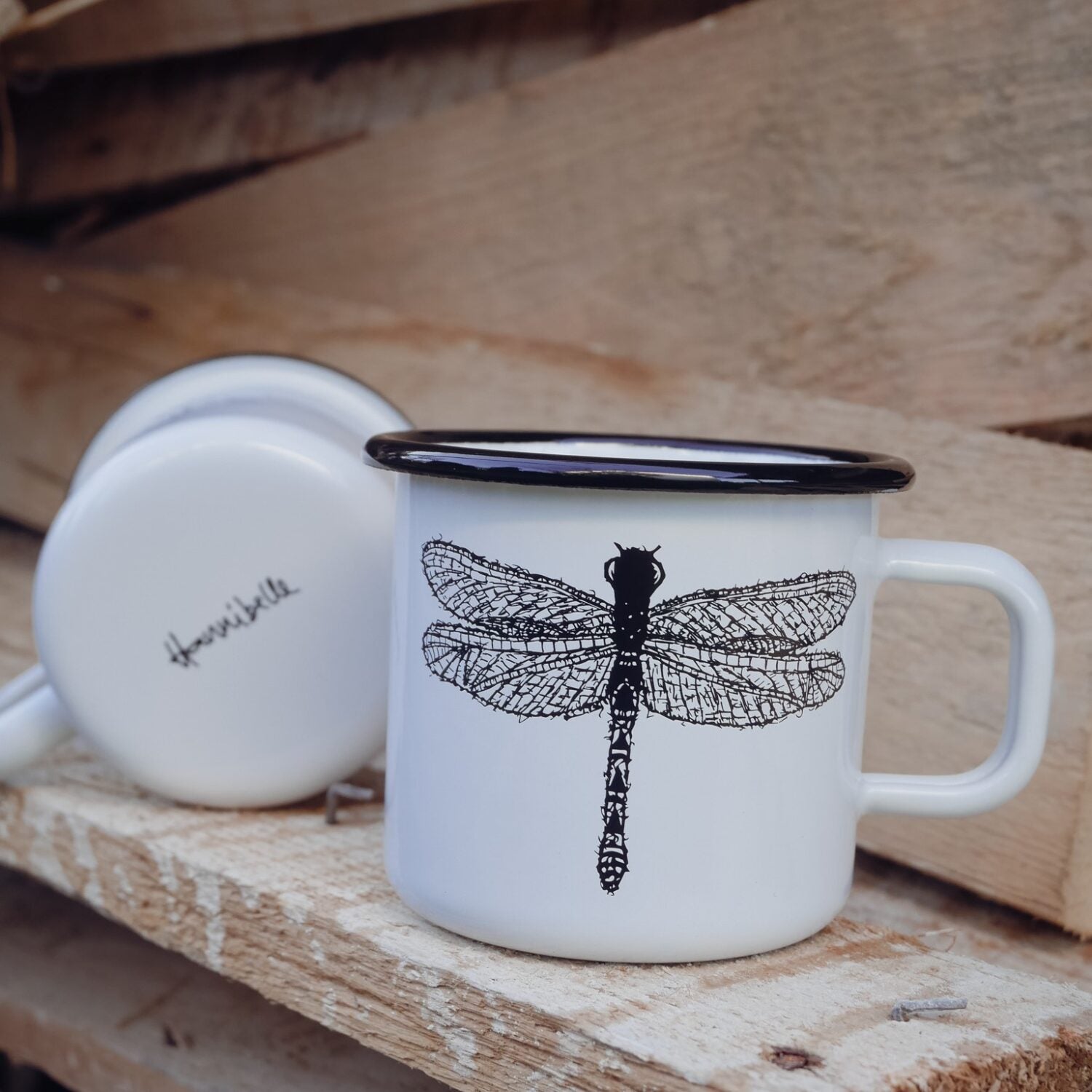 Weiße Emaille-Tasse mit einer Libellenzeichnung und schwarzem Rand, daneben eine umgedrehte Tasse mit Schriftzug.