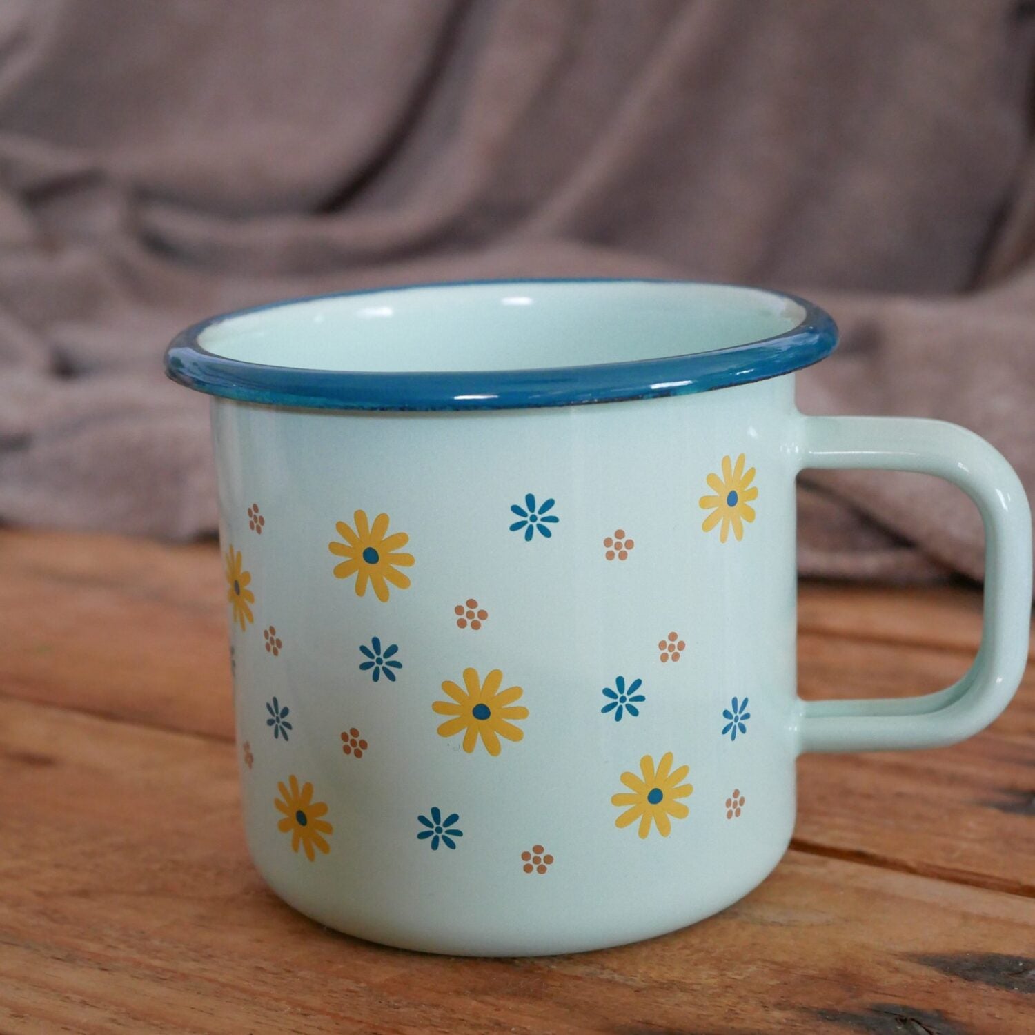 Hellgrüner Emaillebecher mit gelbem und blauem Blumenmuster, auf einem Holztisch vor einer weichen Decke.