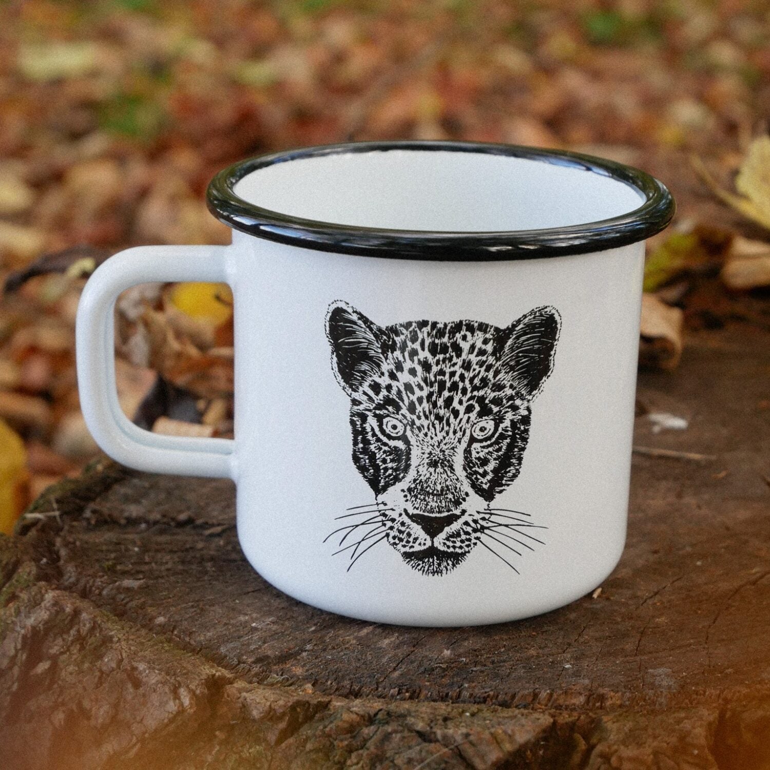 Weiße Tasse mit Leopardenkopf-Illustration steht auf einem Baumstumpf im Wald, umgeben von Herbstlaub.