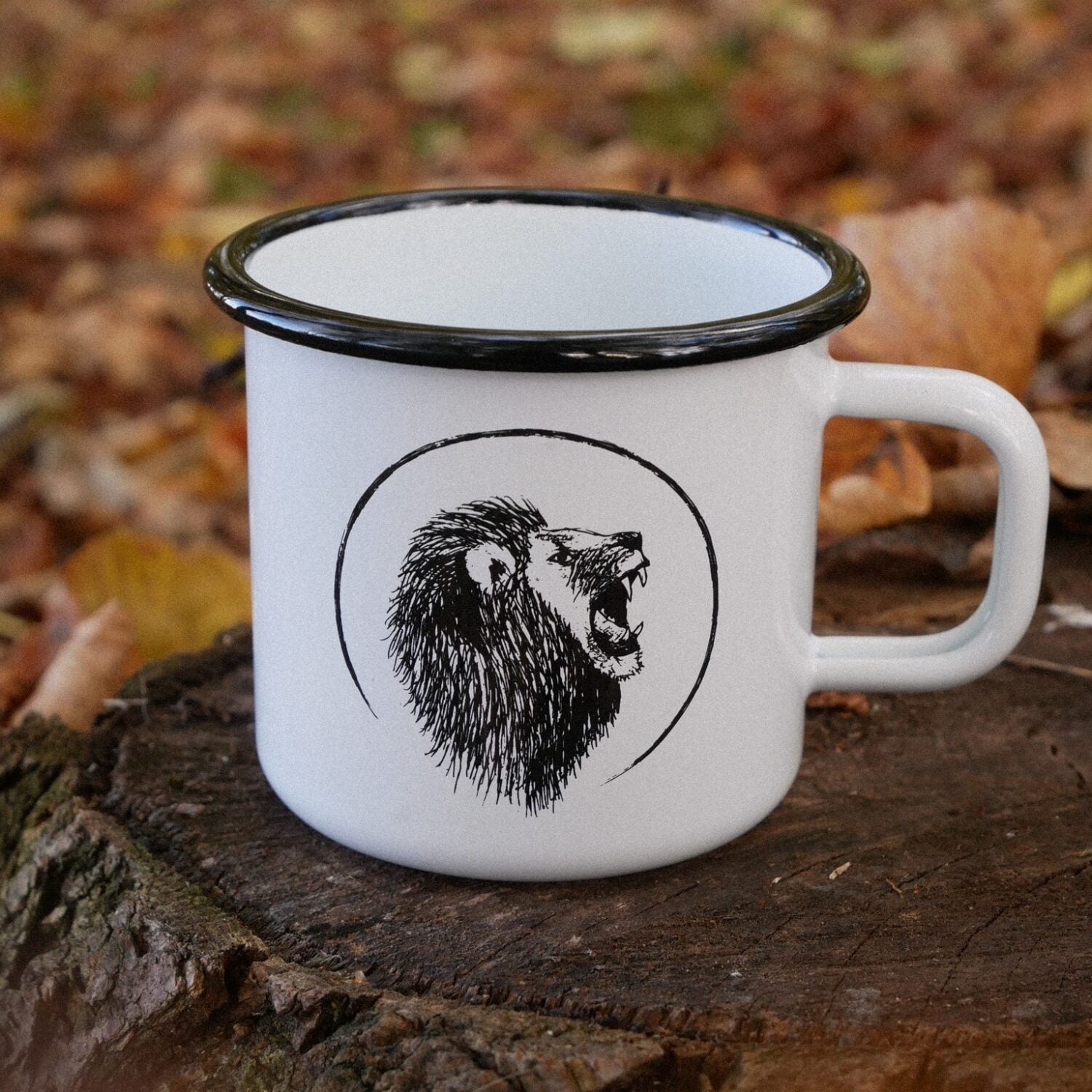 Weißer Emaillebecher mit schwarzem Löwenmotiv, auf einem Baumstumpf sitzend, umgeben von Herbstblättern.