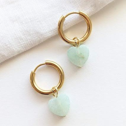 Goldene Creolen mit anhängenden, hellblauen Herzsteinen auf hellem Hintergrund.