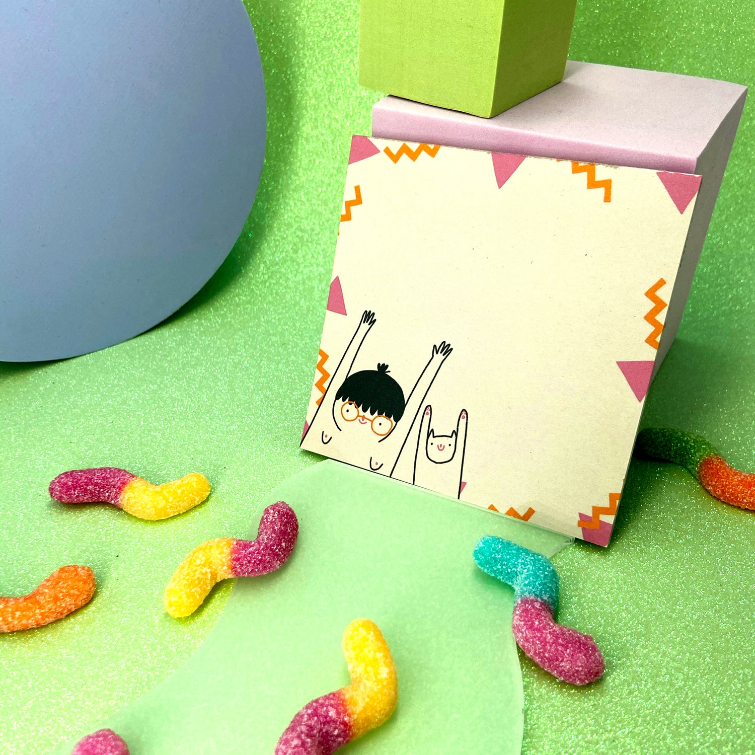 Bunte Gummiwürmer auf grünem Hintergrund, neben einer dekorativen Karte mit niedlicher Zeichnung.