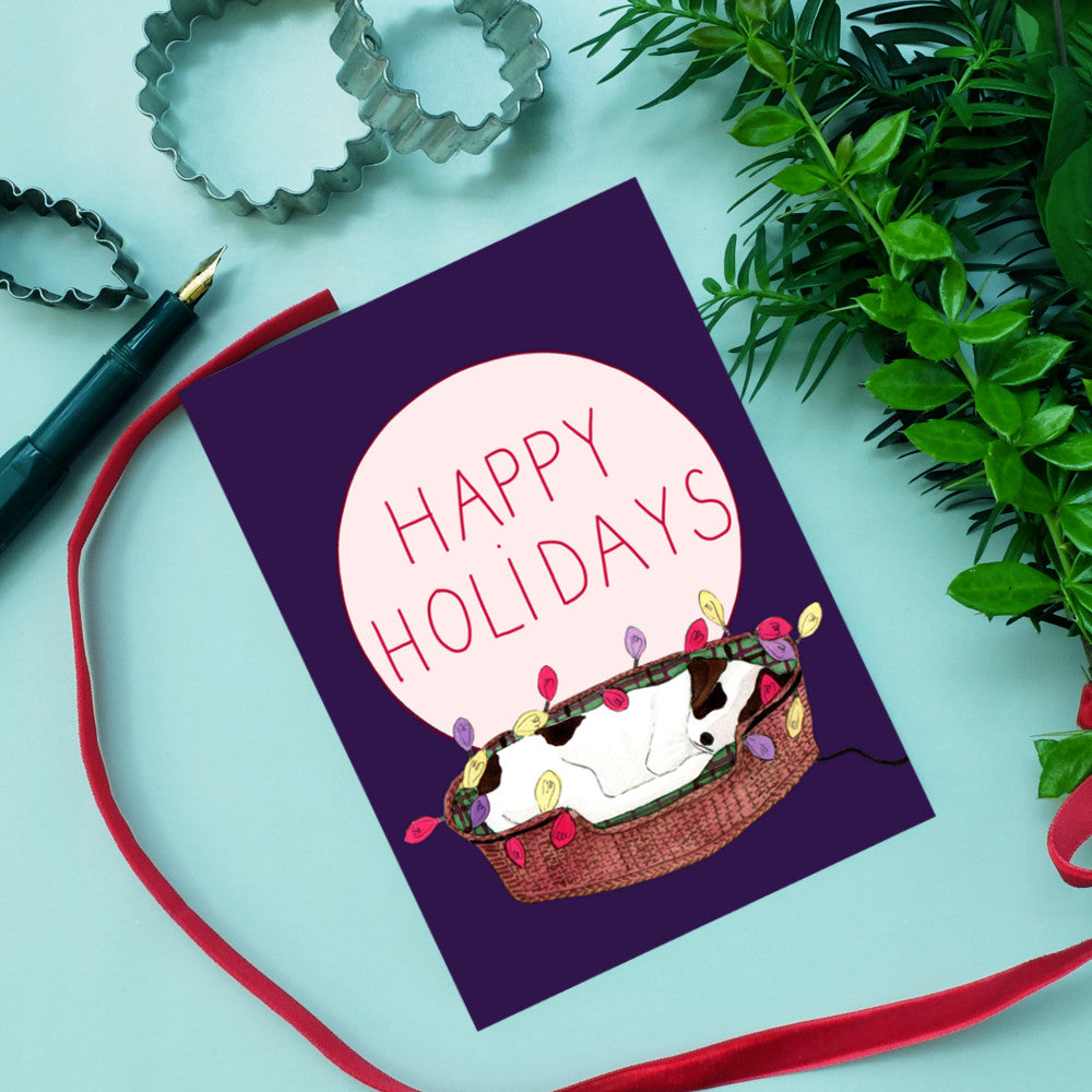 Urlaubskarte mit einem Hund in einem Korb, geschmückt mit bunten Lichtern und dem Text "Happy Holidays".