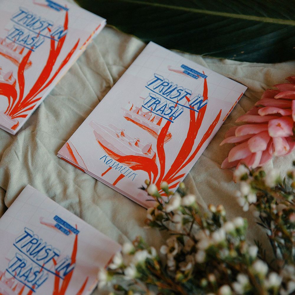 Magazine mit der Aufschrift "TRUST IN TRASH" liegt auf Stoff, umgeben von Blumen und Blättern.