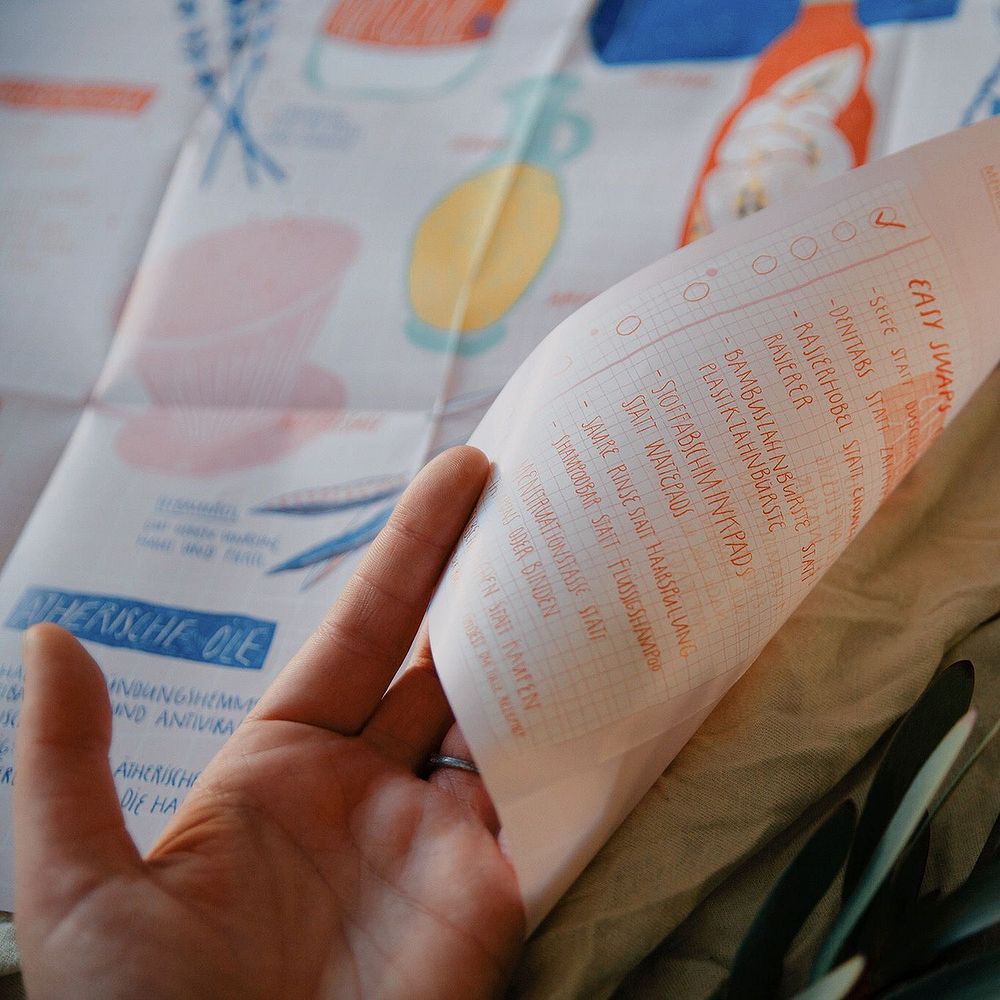 Eine Hand hält eine gefaltete Liste mit bunten Illustrationen und handgeschriebenem Text.