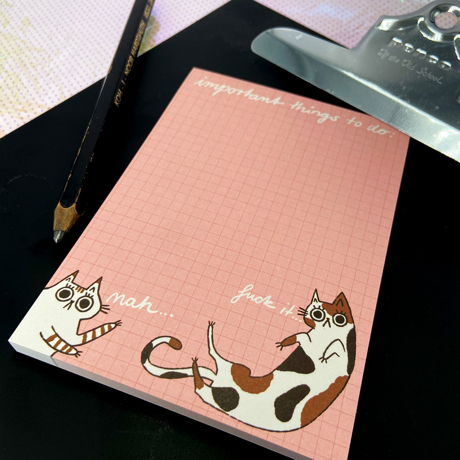Ein rosa Notizblock mit karierter Struktur zeigt zwei gezeichnete Katzen mit humorvollen Aussagen.