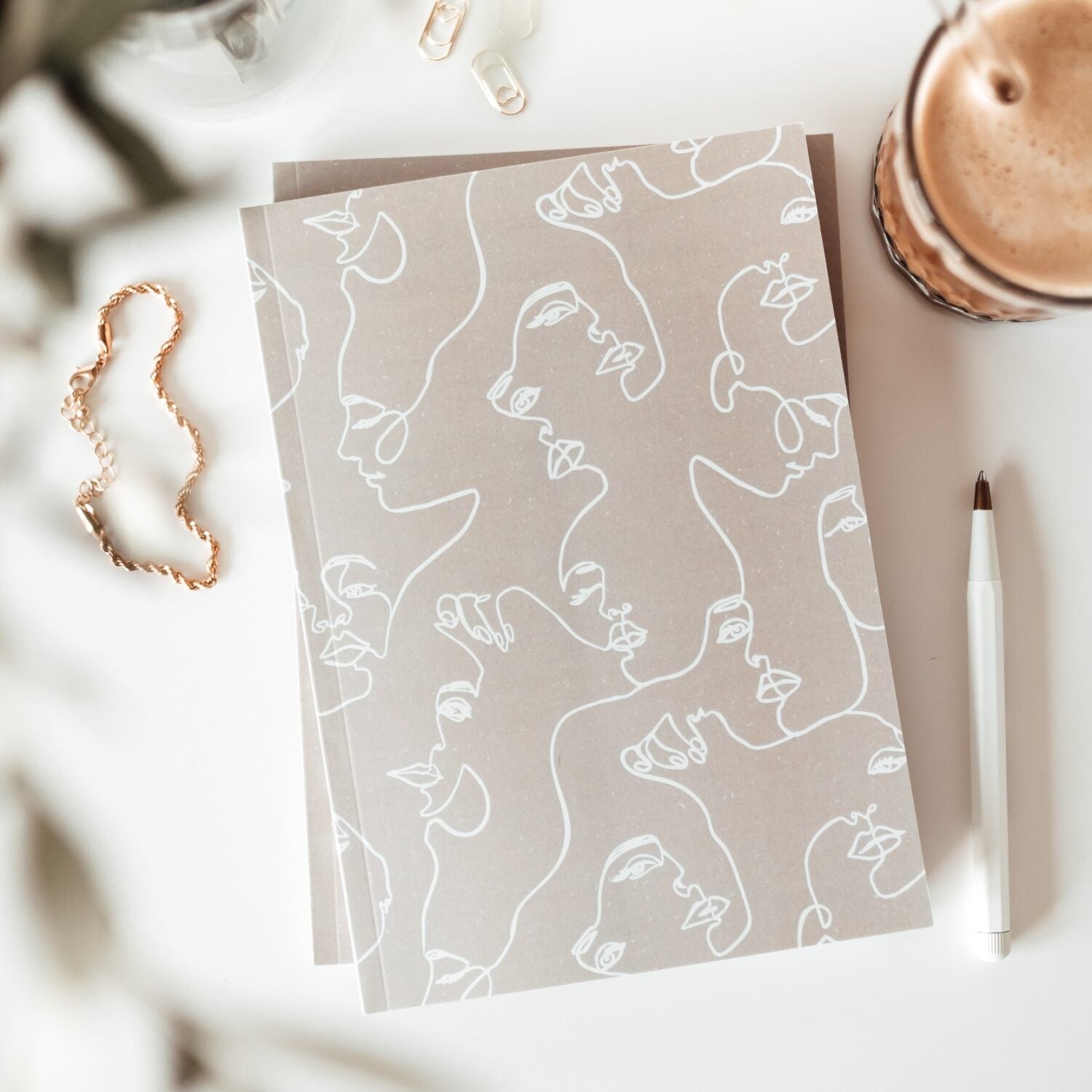 Notizbücher mit abstrakten Gesichtern, ein Stift, ein Getränk und eine goldene Kette auf einem weißen Tisch.