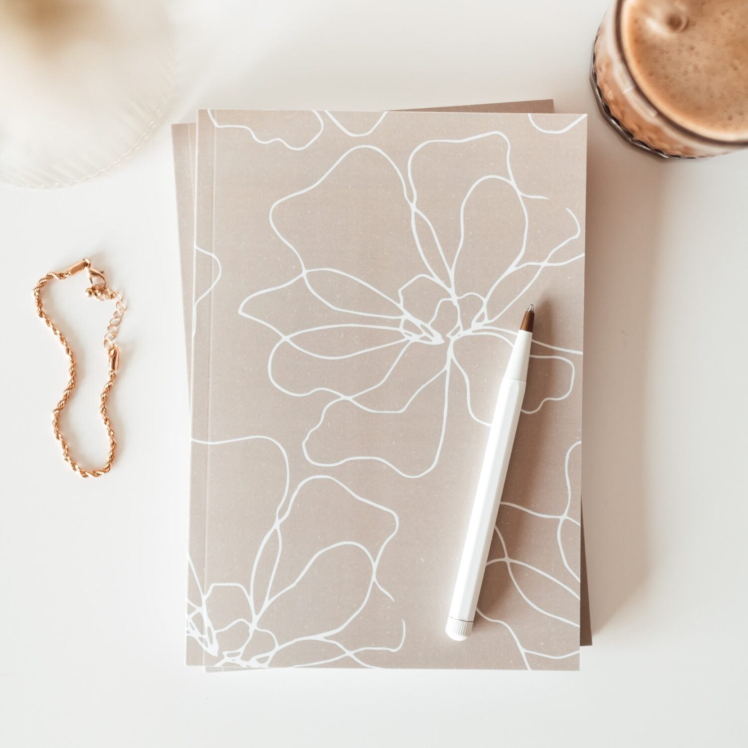 Notizbuch mit Blumendesign, weißem Stift und goldfarbenem Armband auf einem Tisch neben einem Glas mit Latte.