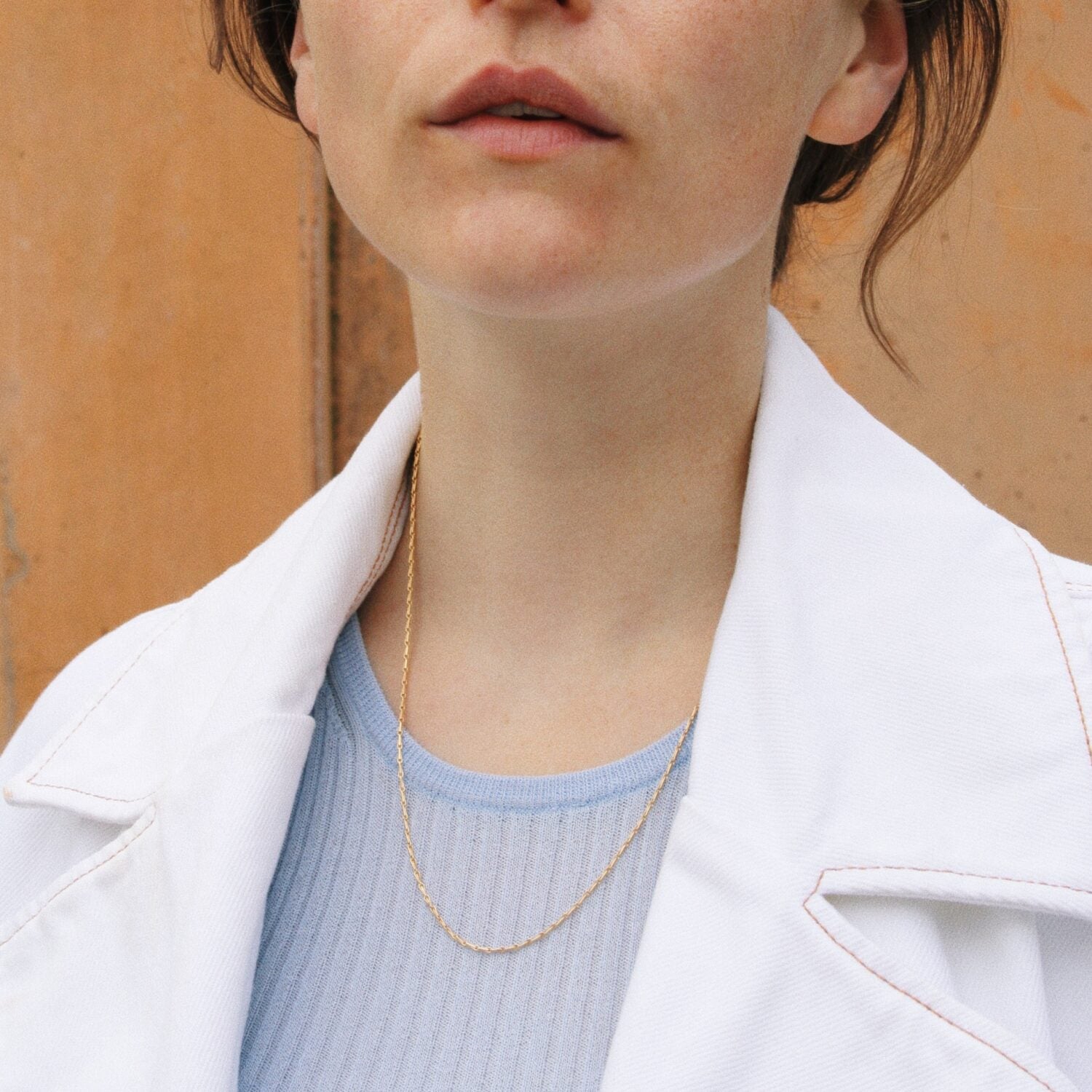 Frau in weißer Jacke und blauem Oberteil trägt eine feine goldene Halskette vor einem orangefarbenen Hintergrund.