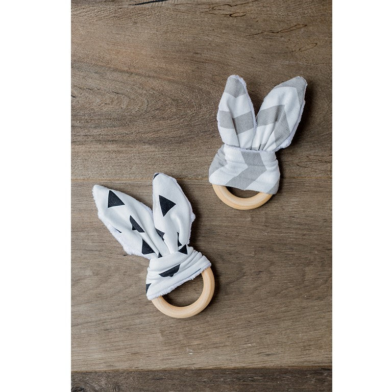 Zwei Beißringe mit Hasenohren auf Holzuntergrund, einer mit Dreiecken, der andere mit Streifenmuster.