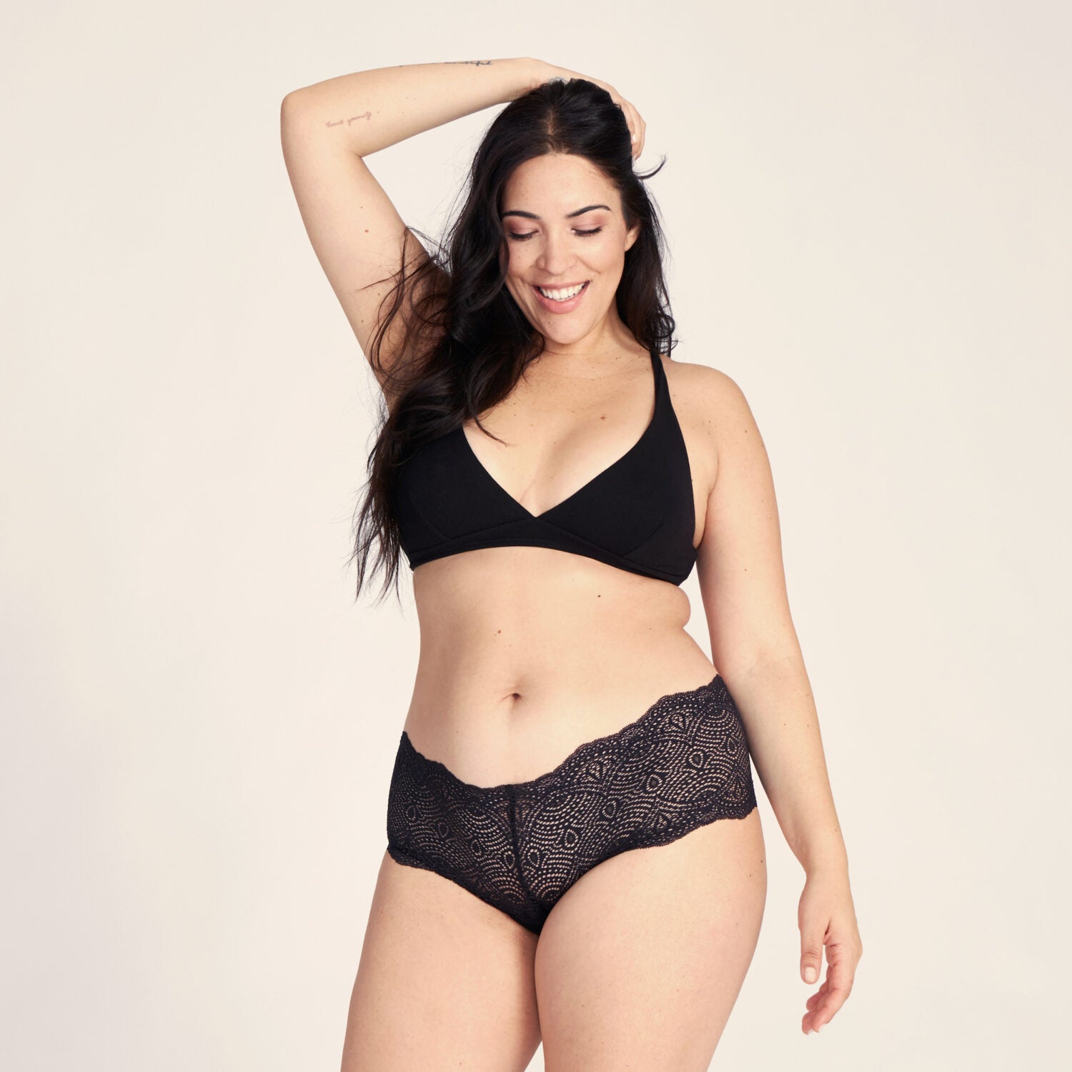 Frau mit langen Haaren trägt schwarzen BH und Spitzenhöschen, lächelnd und mit einer Hand im Haar.