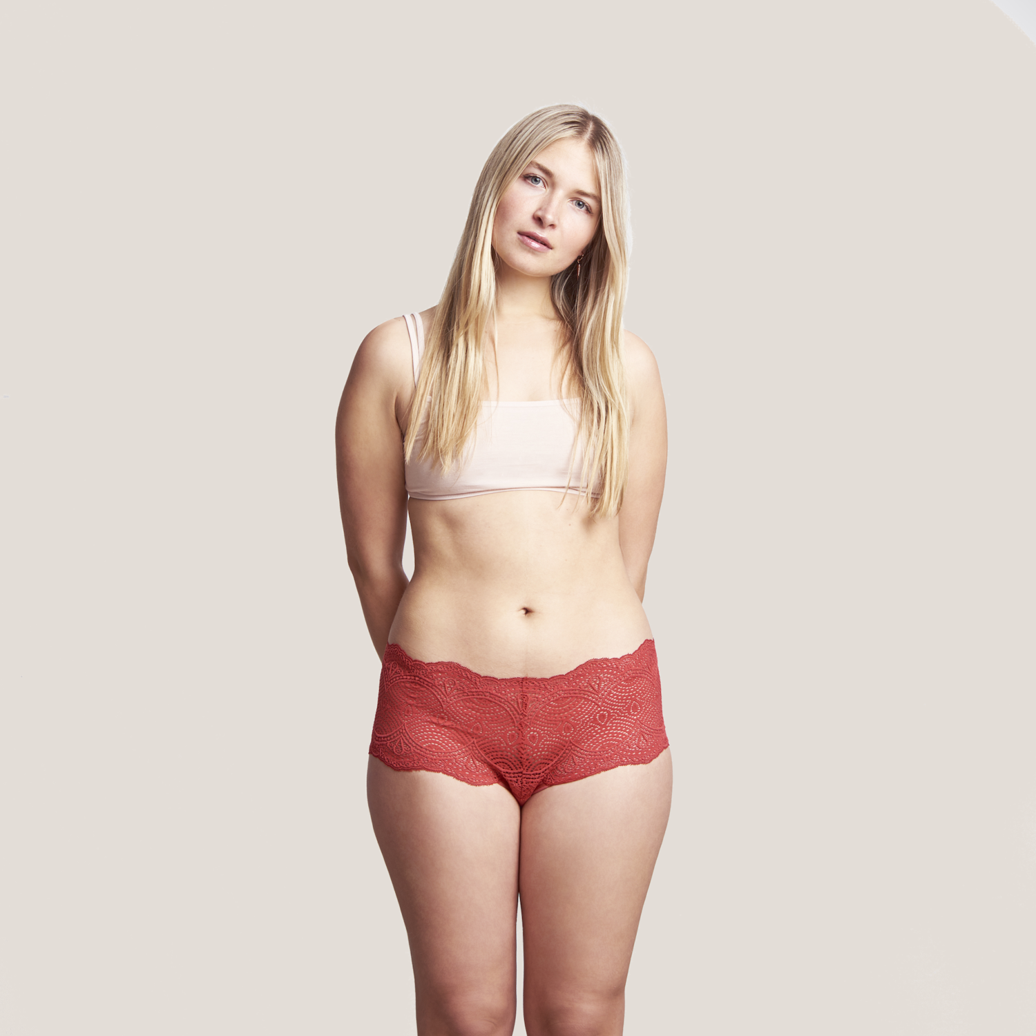 Eine Frau in Unterwäsche steht vor einem neutralen Hintergrund, trägt ein beiges Oberteil und rote Spitzenhöschen.