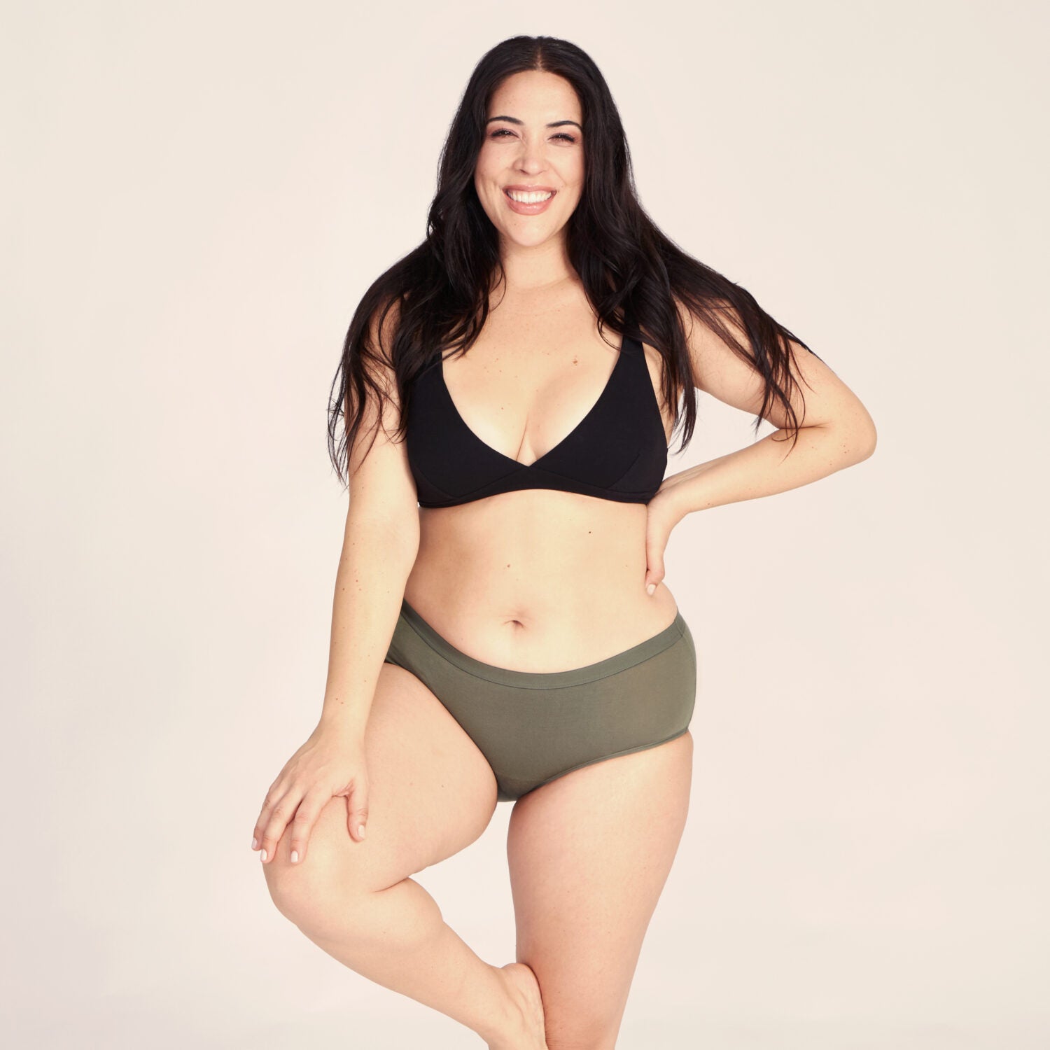 Frau mit schwarzem Oberteil und grüner Unterhose lächelt und steht auf einem Bein gegen einen hellen Hintergrund.