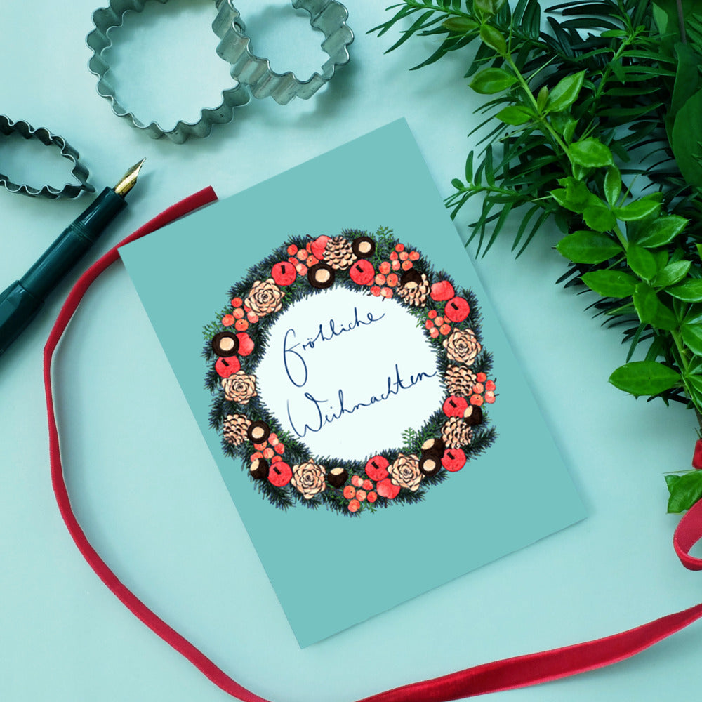 Weihnachtskarte mit dekorativem Kranz und "Fröhliche Weihnachten" in der Mitte, umgeben von grünen Blättern und rotem Band.