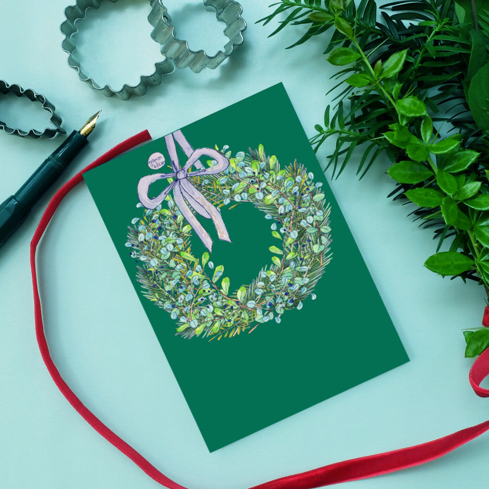 Grüne Grußkarte mit Kranz und Schleife, umgeben von Keksausstechern, Stift, rotem Band und Pflanzen.