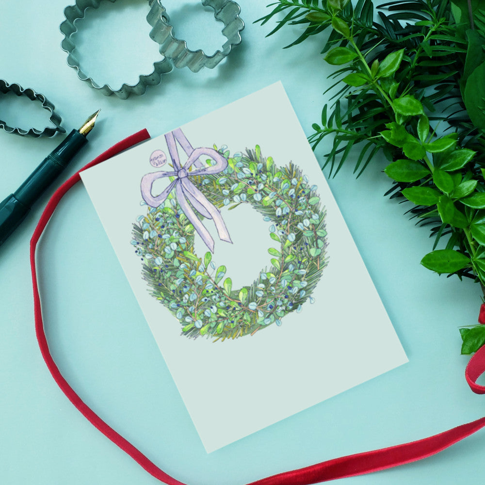 Aquarellkarte mit Kranz und Schleife, umgeben von Plätzchenausstechern, Stift, Blattwerk und rotem Band auf hellblauem Hintergrund.