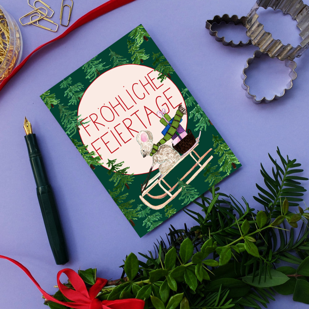 Weihnachtskarte mit einer Maus auf einem Schlitten und der Aufschrift "Fröhliche Feiertage" umgeben von Tannenzweigen.