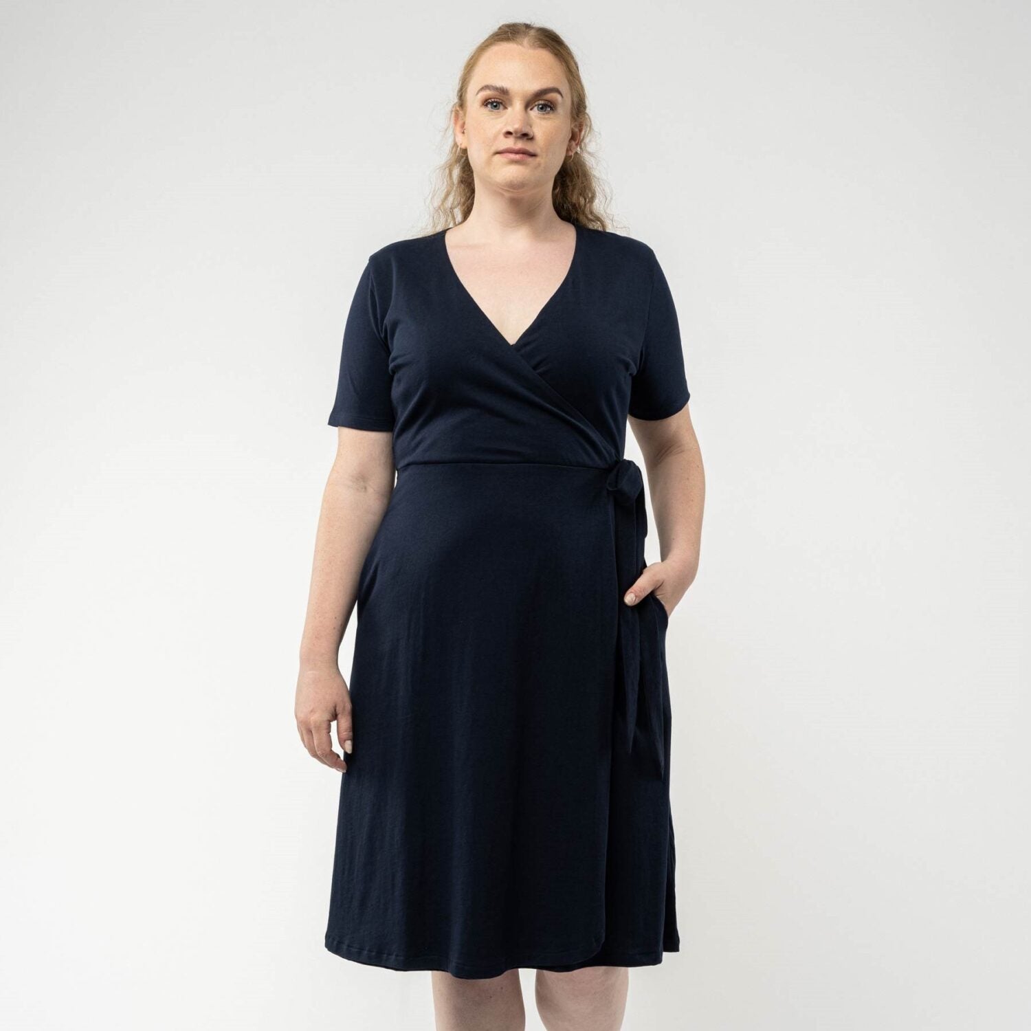 Frau in einem dunkelblauen Wickelkleid mit kurzen Ärmeln, Hand in der Tasche, vor hellem Hintergrund.