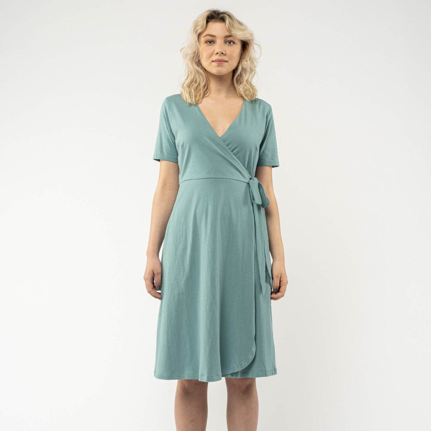 Frau in einem türkisfarbenen Wickelkleid steht vor einem weißen Hintergrund.