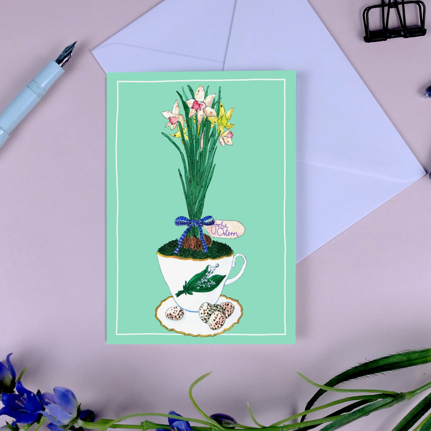 Illustrierte Karte mit Narzissen in einer Teetasse, umgeben von Ostereiern, auf einem Tisch mit Umschlag und Stift.