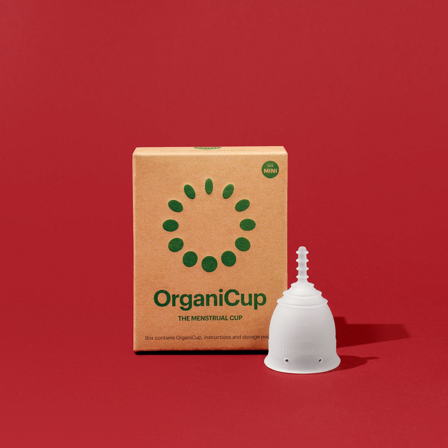 Pappschachtel der OrganiCup Menstruationstasse neben einer weißen Menstruationstasse vor rotem Hintergrund.