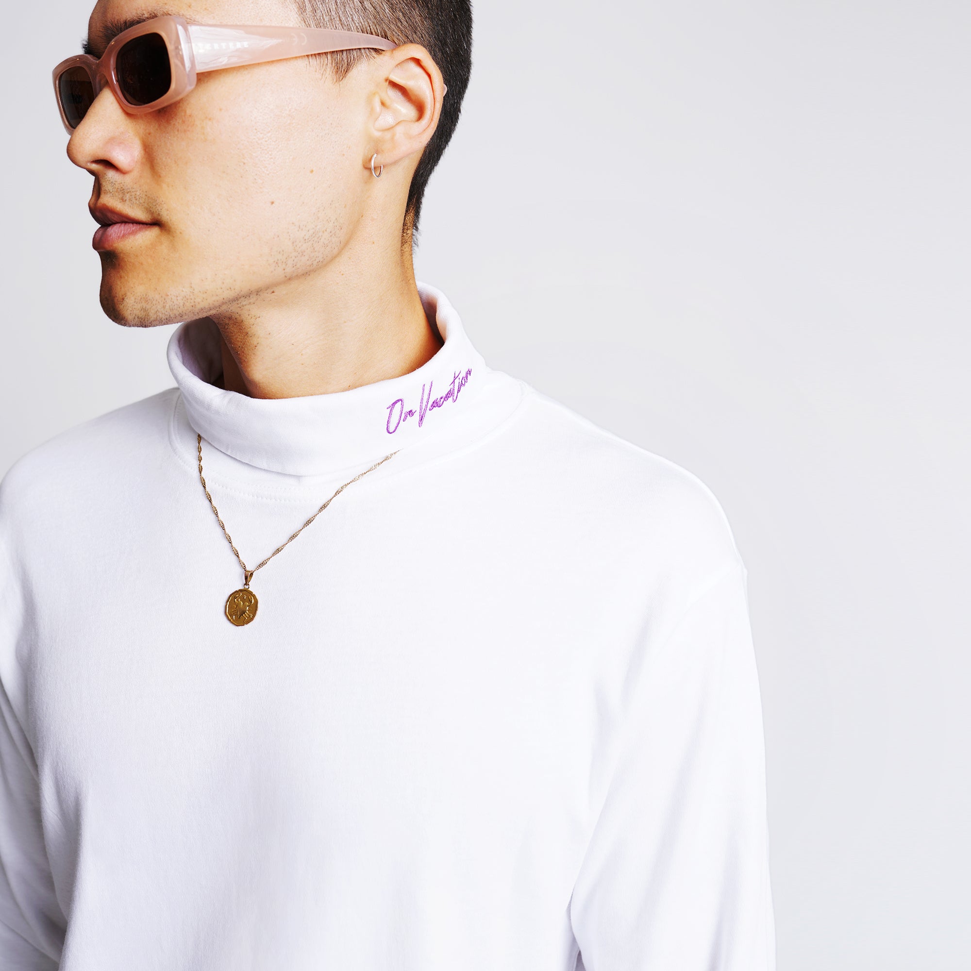 Person mit Kurzhaarschnitt trägt Sonnenbrille, weißen Rollkragenpullover mit Aufschrift "On Vacation" und eine Halskette.