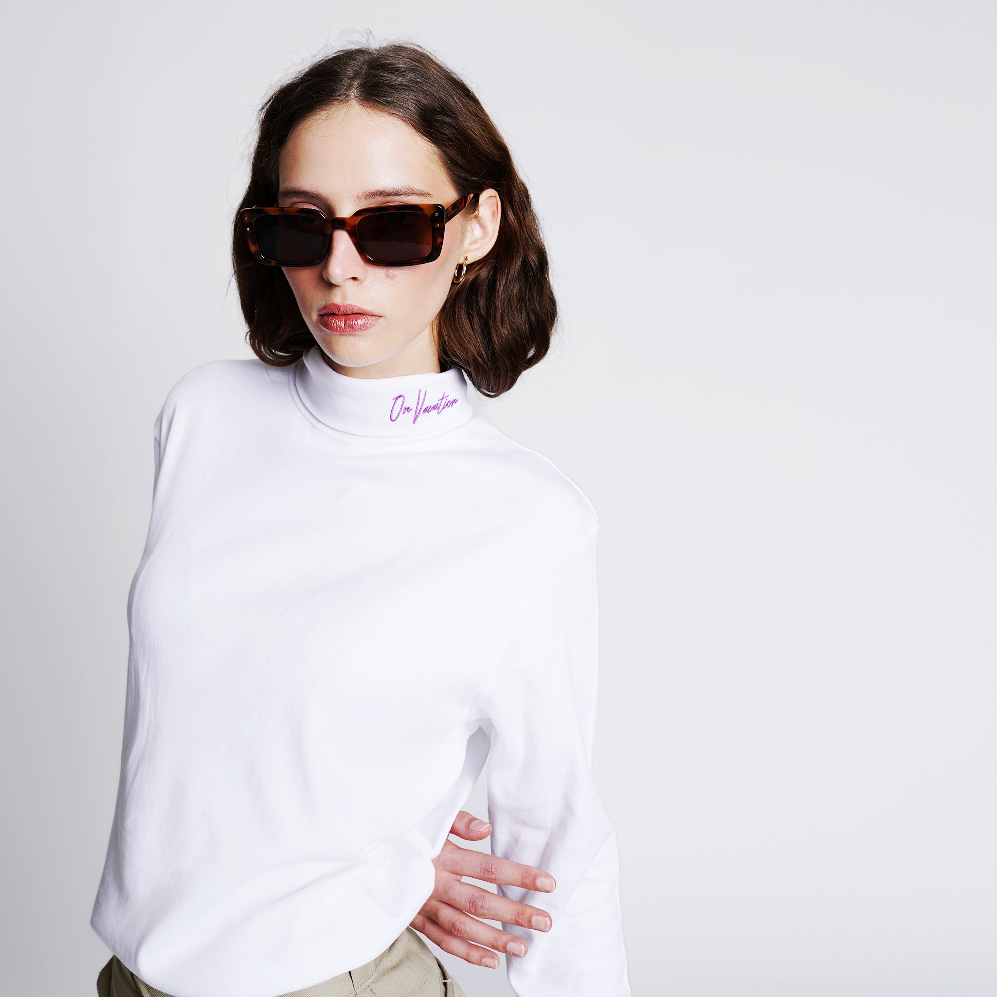 Frau mit braunem Haar trägt eine Sonnenbrille und einen weißen Pullover mit Aufschrift „On Vacation“ am Kragen.