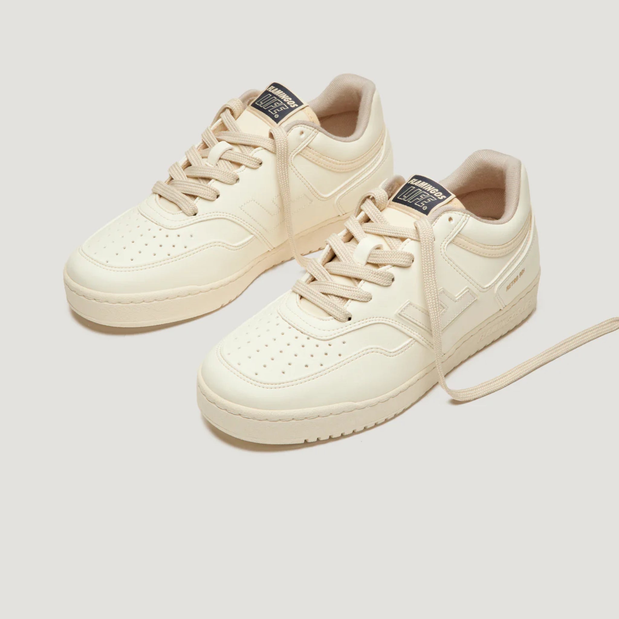 Beige Sneaker mit Schnürsenkeln, perforiertem Obermaterial und dunklem Logo auf der Zunge, auf hellem Hintergrund.