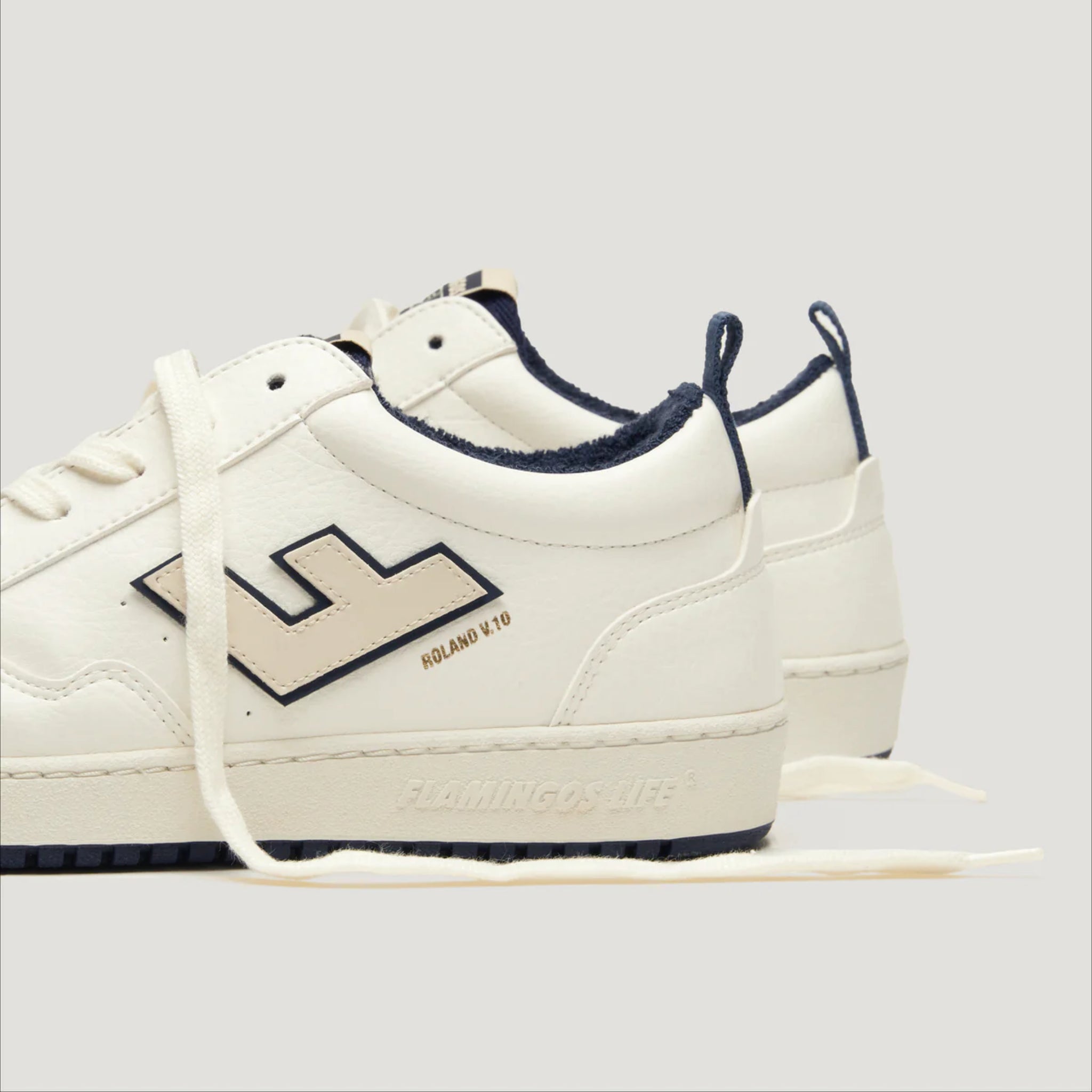 Weiße Sneaker mit dunkelblauen Details und seitlichem Logo auf einer hellgrauen Oberfläche.