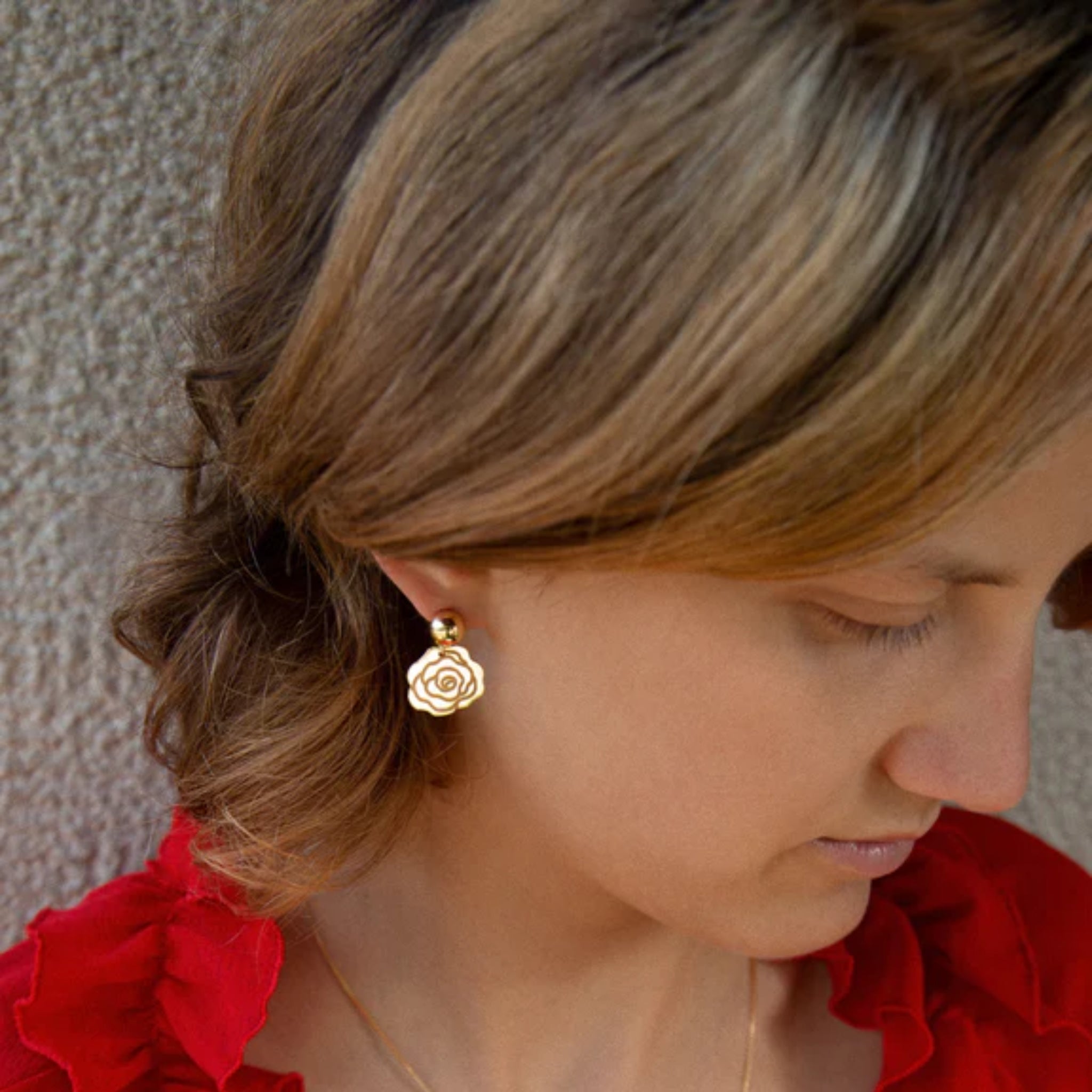 Frau mit seitlich gescheiteltem Haar trägt einen goldenen Rosenohrring und ein rotes Oberteil vor einer Mauer.