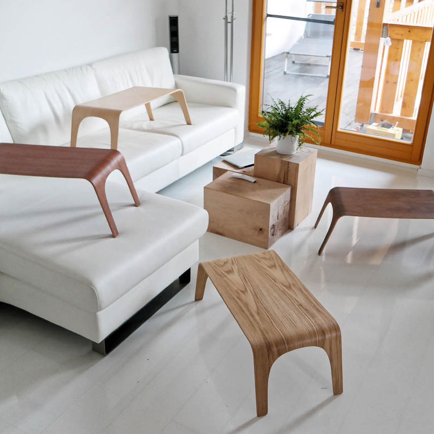 Helles Wohnzimmer mit weißem Sofa, Holzbeistelltischen, Pflanzen und großen Fenstern mit Blick auf einen Balkon.