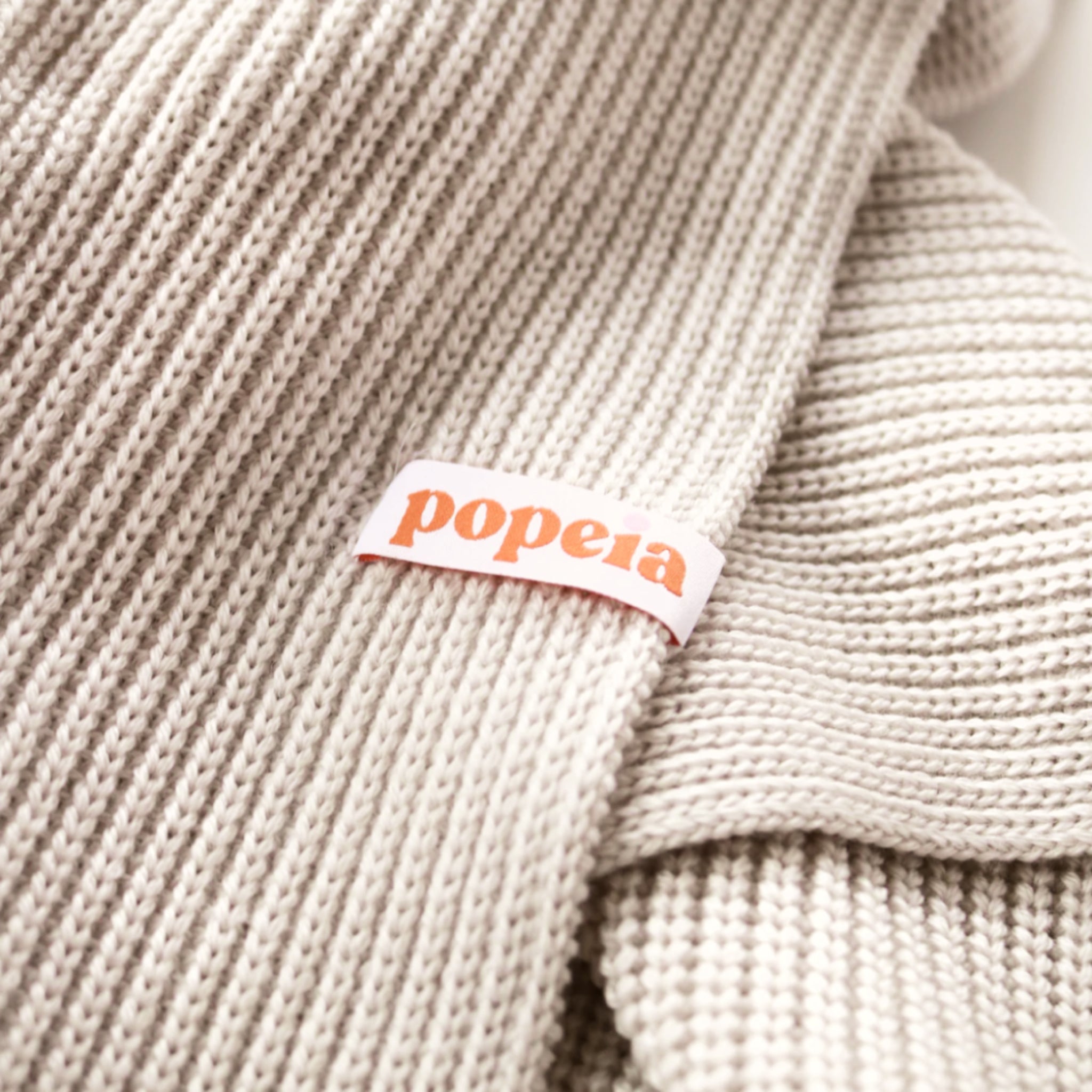 Beigefarbener Strickstoff mit einem Etikett, auf dem "popeia" in orangefarbener Schrift steht.