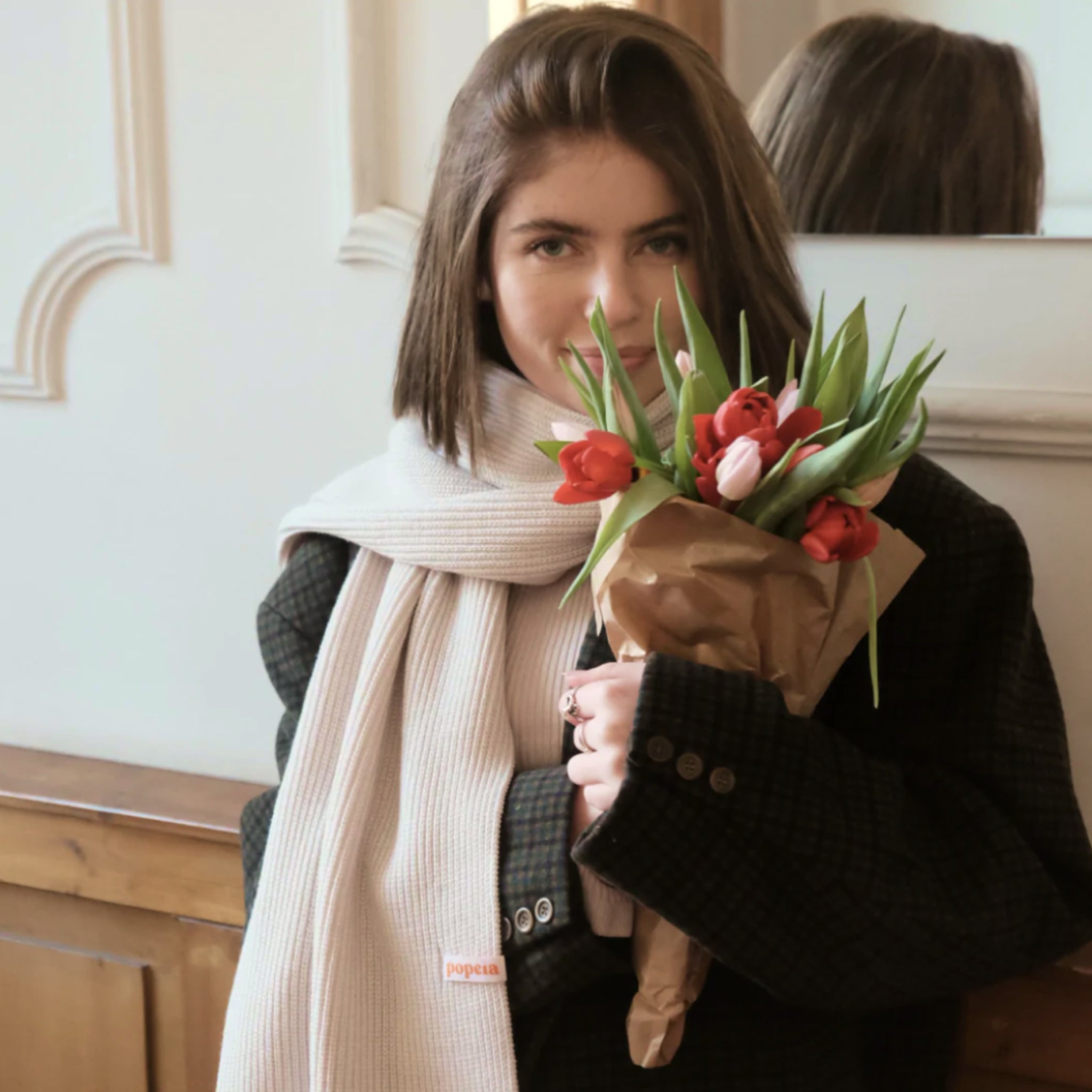 Frau mit weißem Schal und braunem Mantel hält einen Strauß roter und rosa Tulpen vor einem Spiegel.